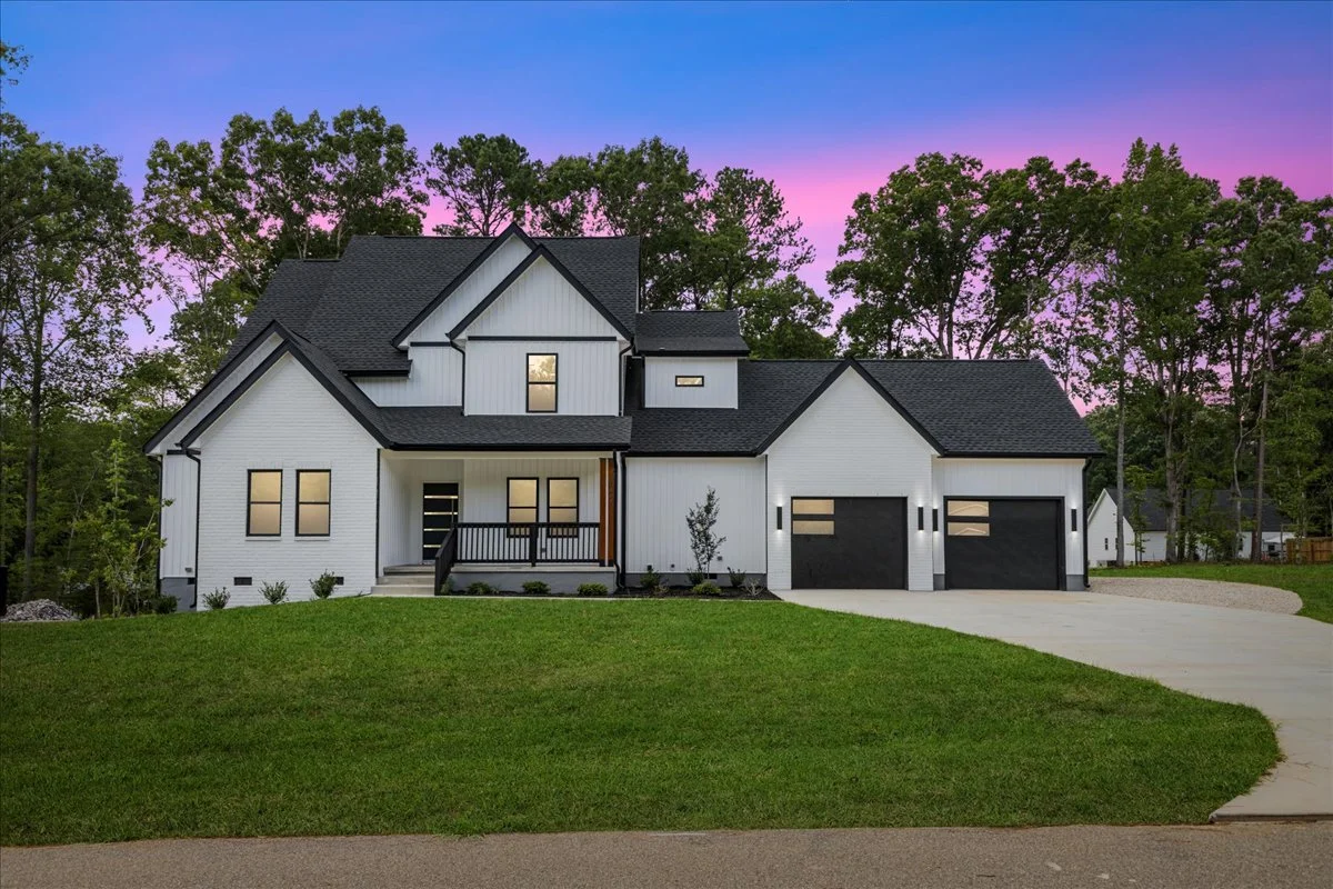 Modern two-story house with white exterior, black roof, and black garage doors, situated on a lawn with trees and a sunset sky in the background.