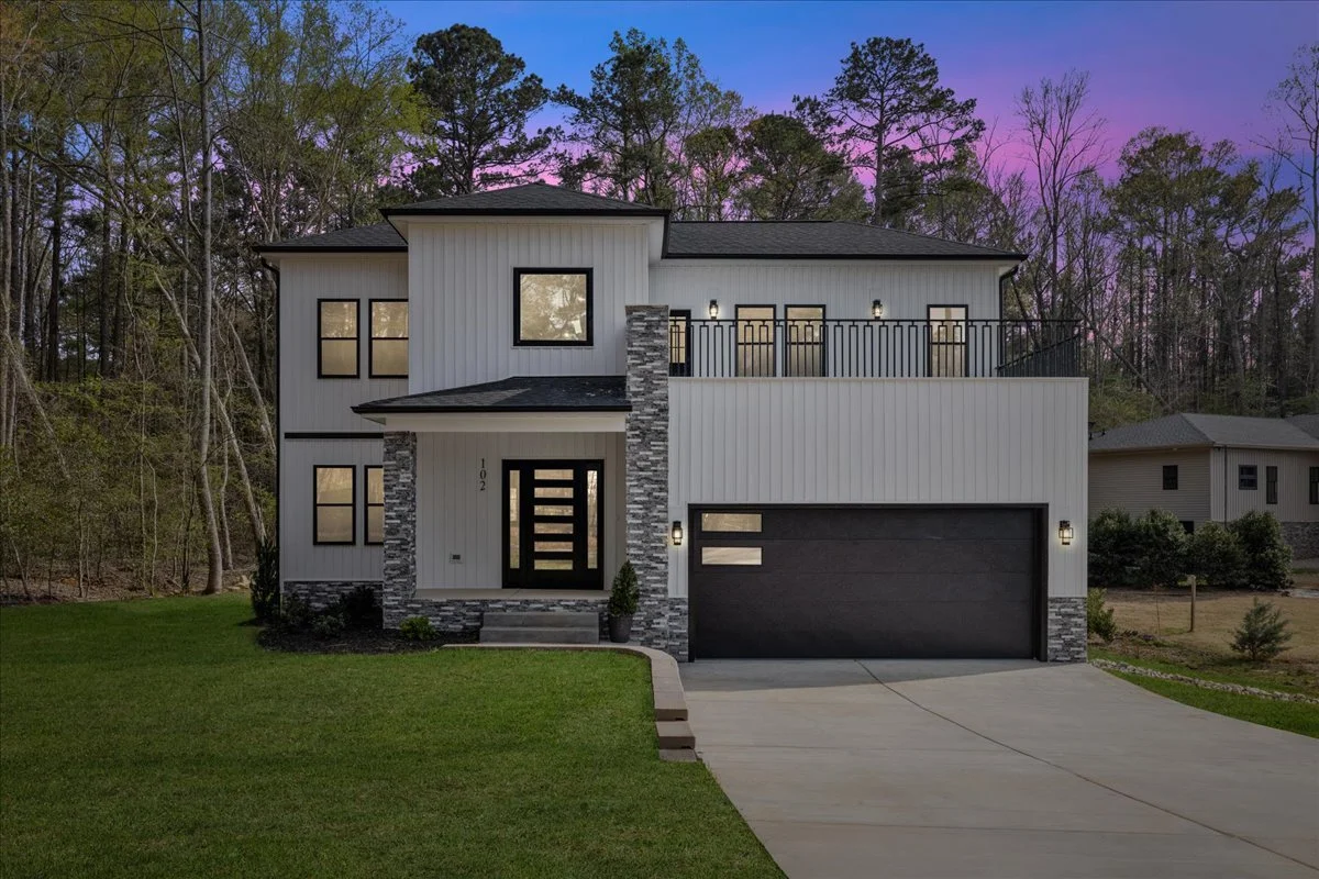 Modern two-story house with gray exterior, black accents, a large attached garage, and a front porch with steps, set against a wooded background at dusk.