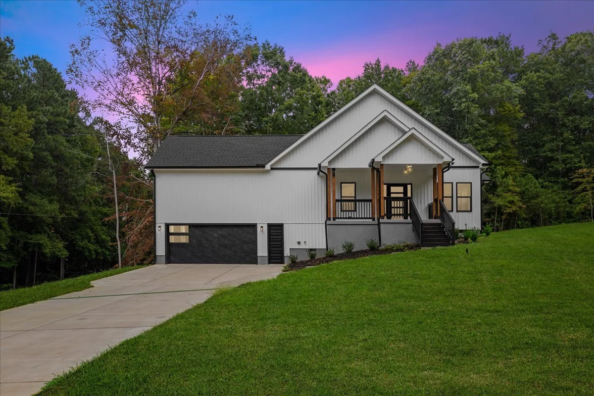A modern house with white exterior siding, black front door, and small front porch with black railing, situated on a grassy hill with a driveway leading to a garage, surrounded by trees and a colorful sunset sky in the background.