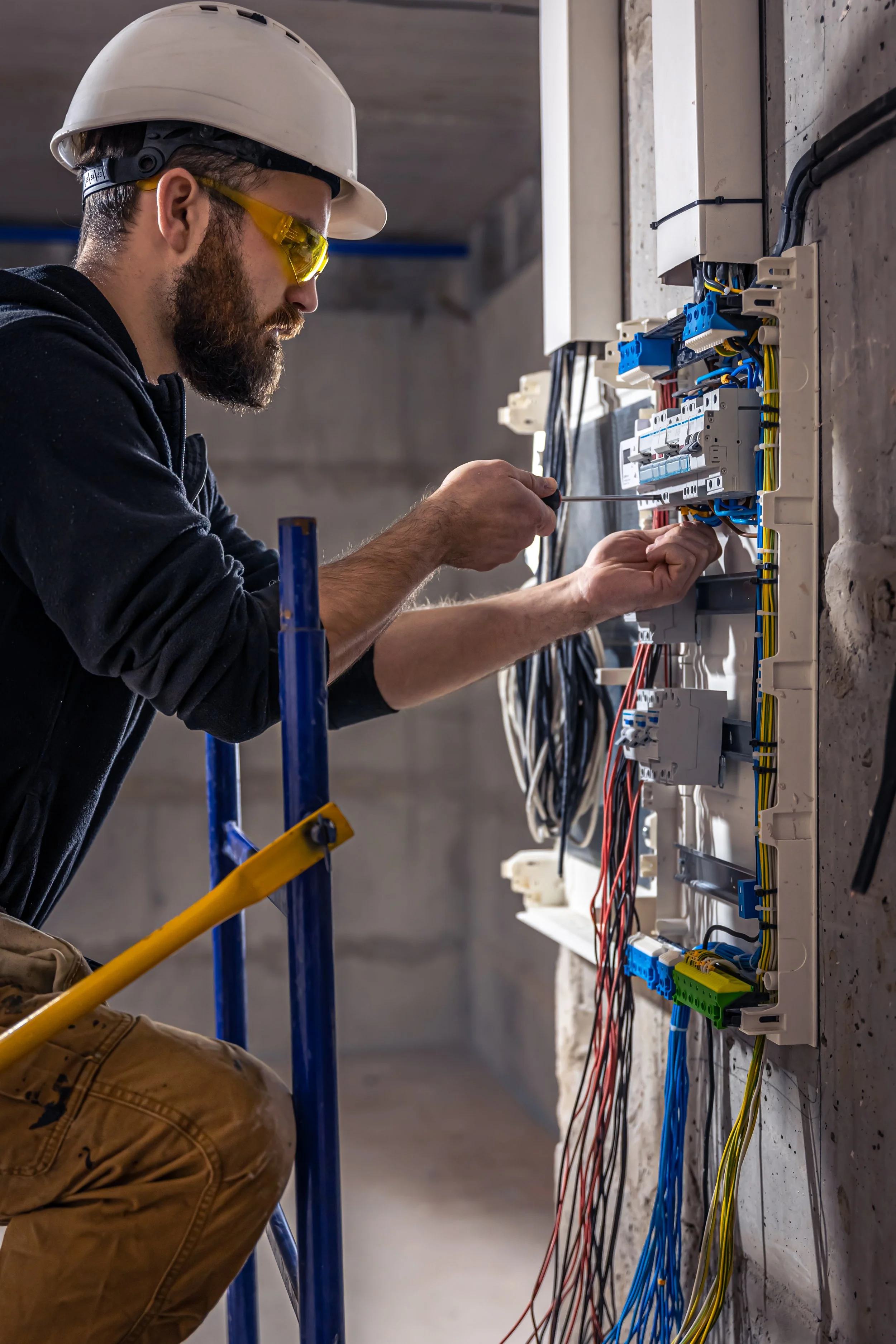 A man wearing a helmet and safety glasses working on an electrical panel with various colored wires.