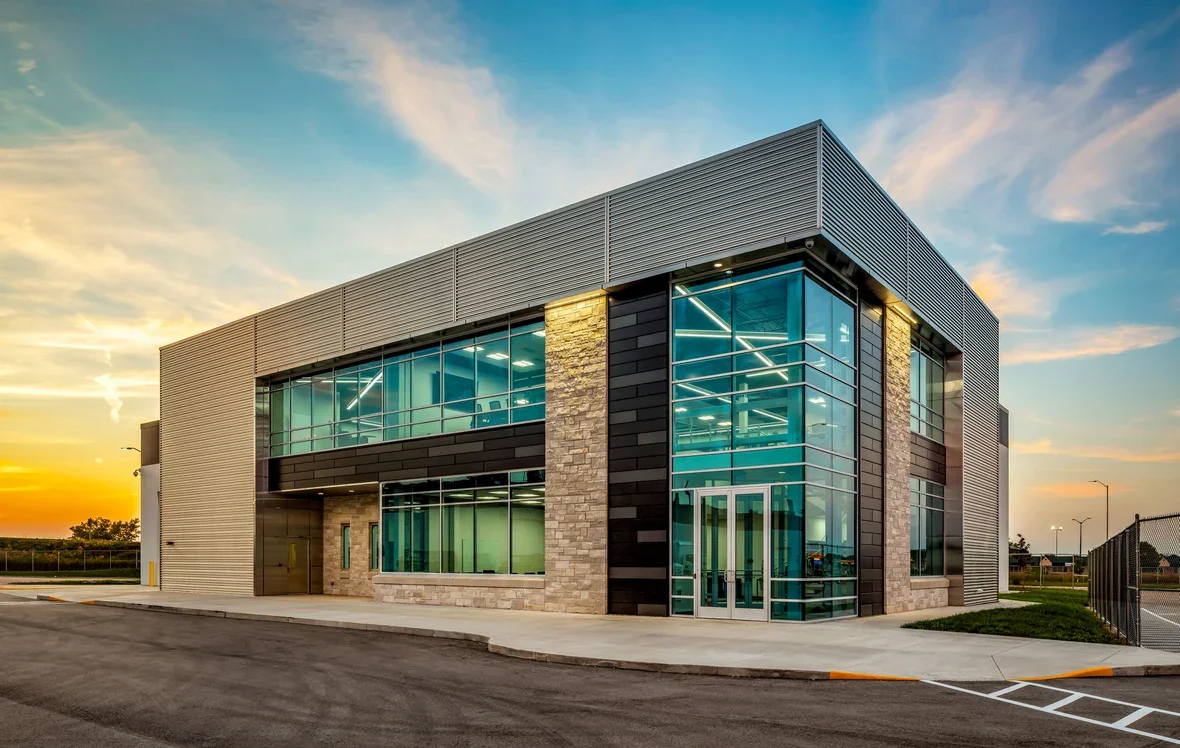Modern two-story commercial building with glass windows, stone and black siding, metal accents, located in a parking lot during sunset.