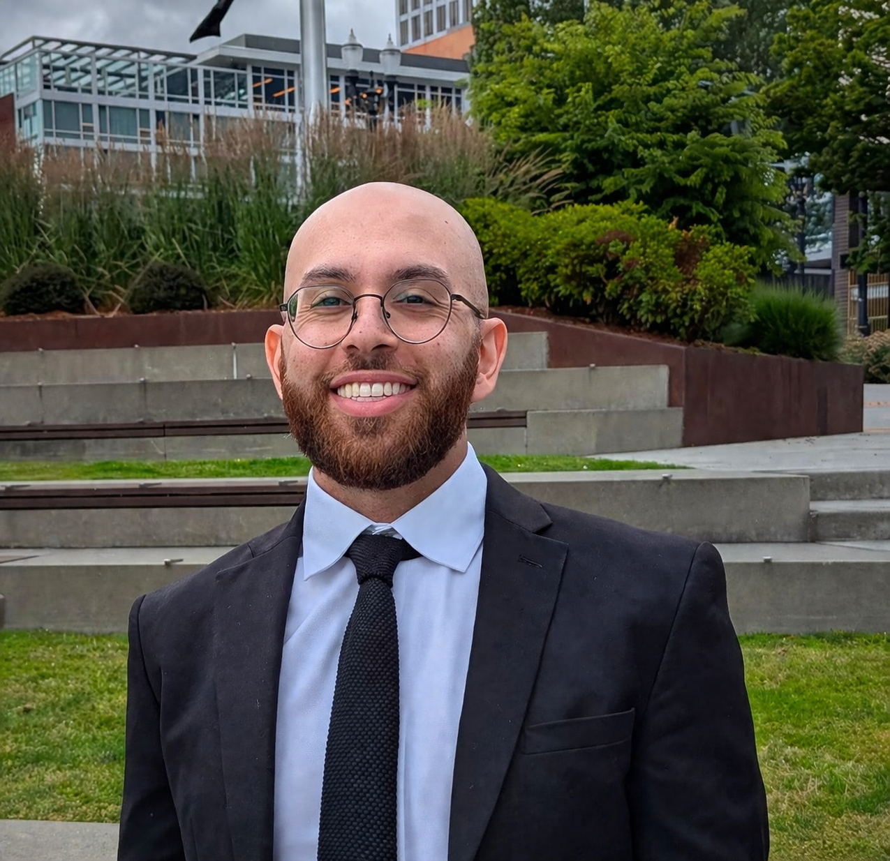 A smiling man with a beard and glasses in a black suit, white shirt, and black tie standing outdoors in front of steps, greenery, and buildings.