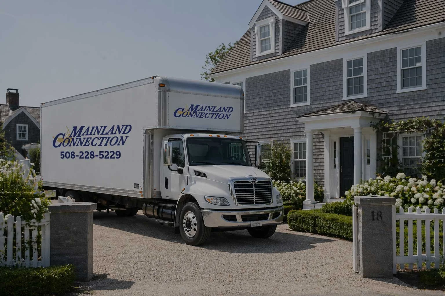 White Nantucket moving truck with 'Mainland Connection' branding parked in front of a house with gray siding and a porch, surrounded by flowering bushes and a white picket fence.