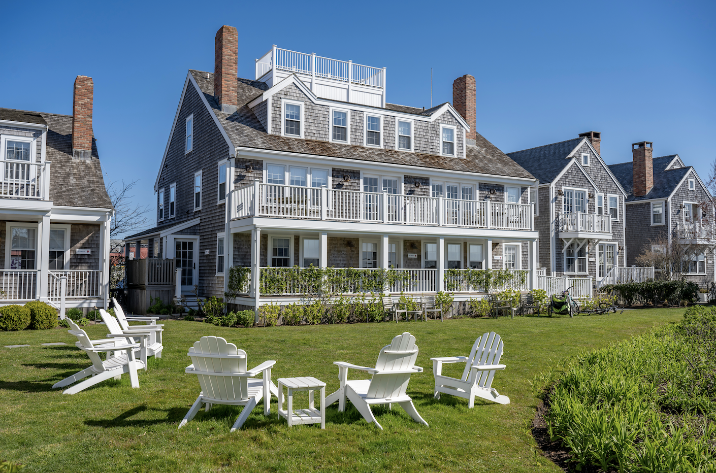 A large multi-story house with gray wooden shingles, white railings, and multiple balconies on a sunny day. There are green bushes in front of the house, and white Adirondack chairs and a small table on the lawn. Bicycle and lawn equipment are visible near the house.