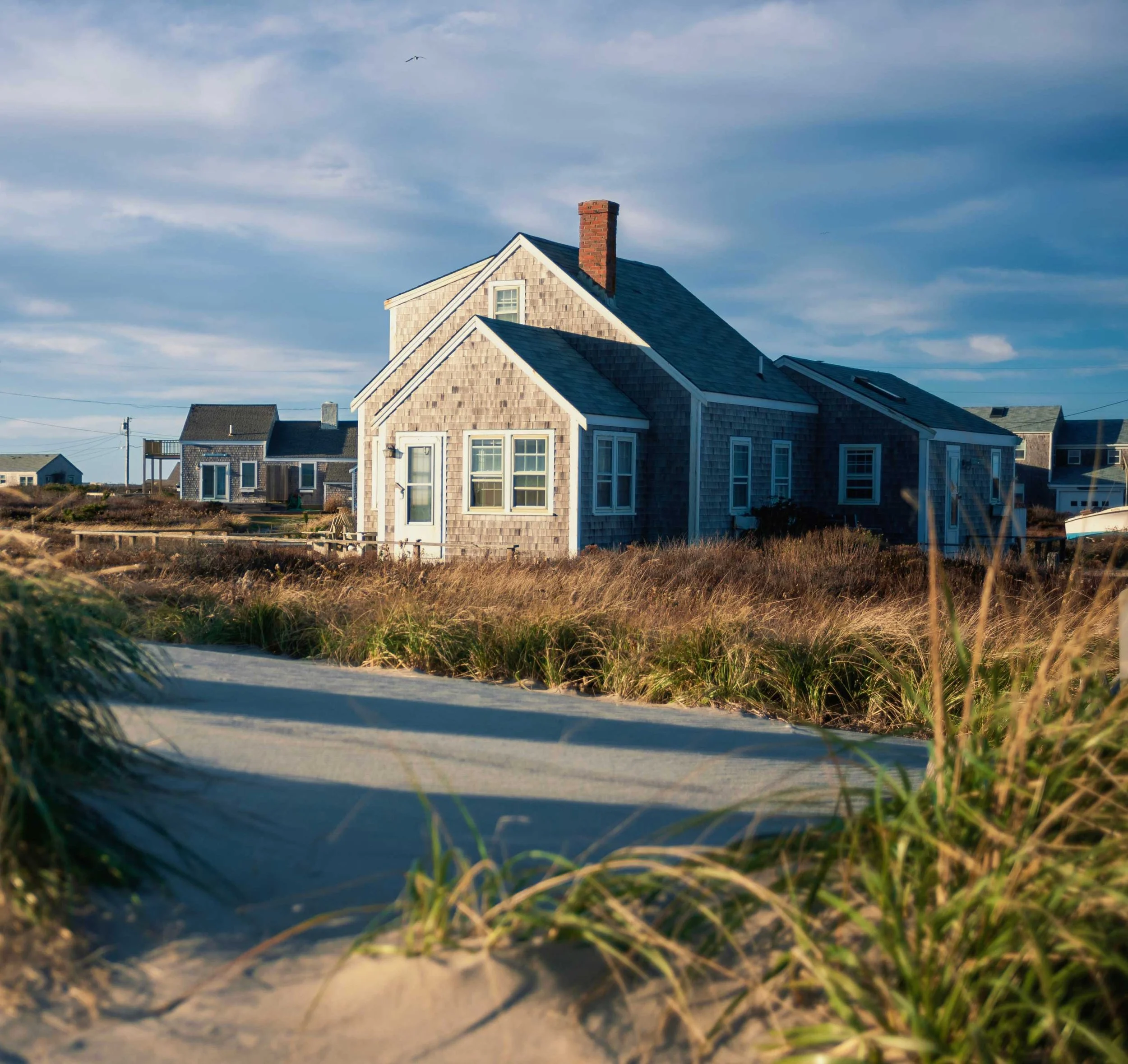 View of sandy dunes with patches of grass and a house with a shingle exterior, located near a blue sky with streaks of white clouds and a bird flying overhead.