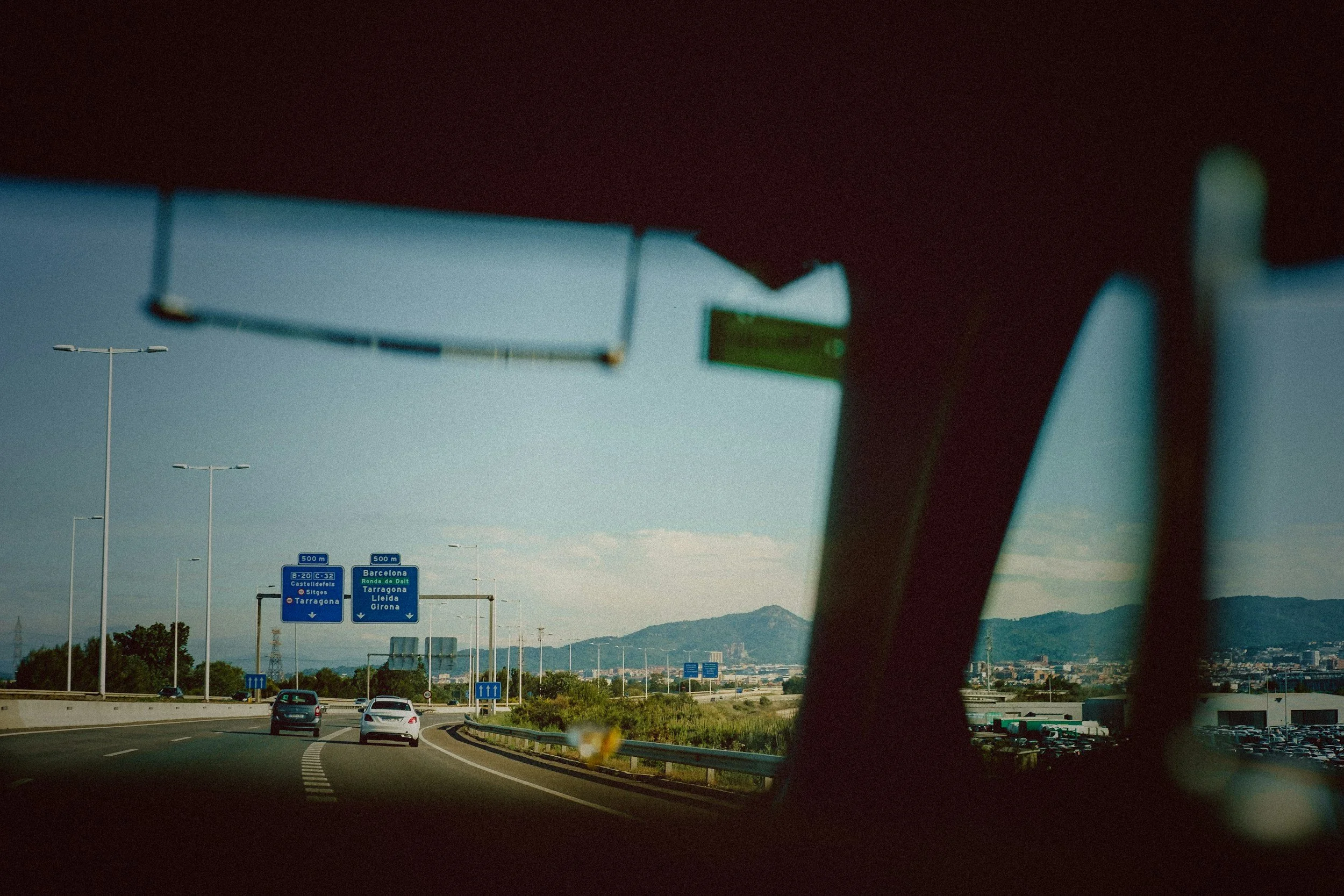 View of a highway through a vehicle's windshield with blue directional road signs indicating routes to Barcelona, Tarragona, Girona, and other locations, with mountains in the background.