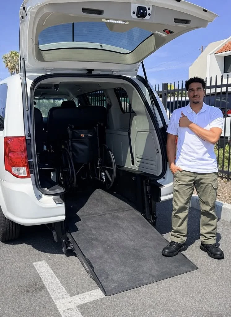 A man stands next to a white van with its rear hatch open, revealing a wheelchair accessible ramp and an interior space designed for wheelchair access.