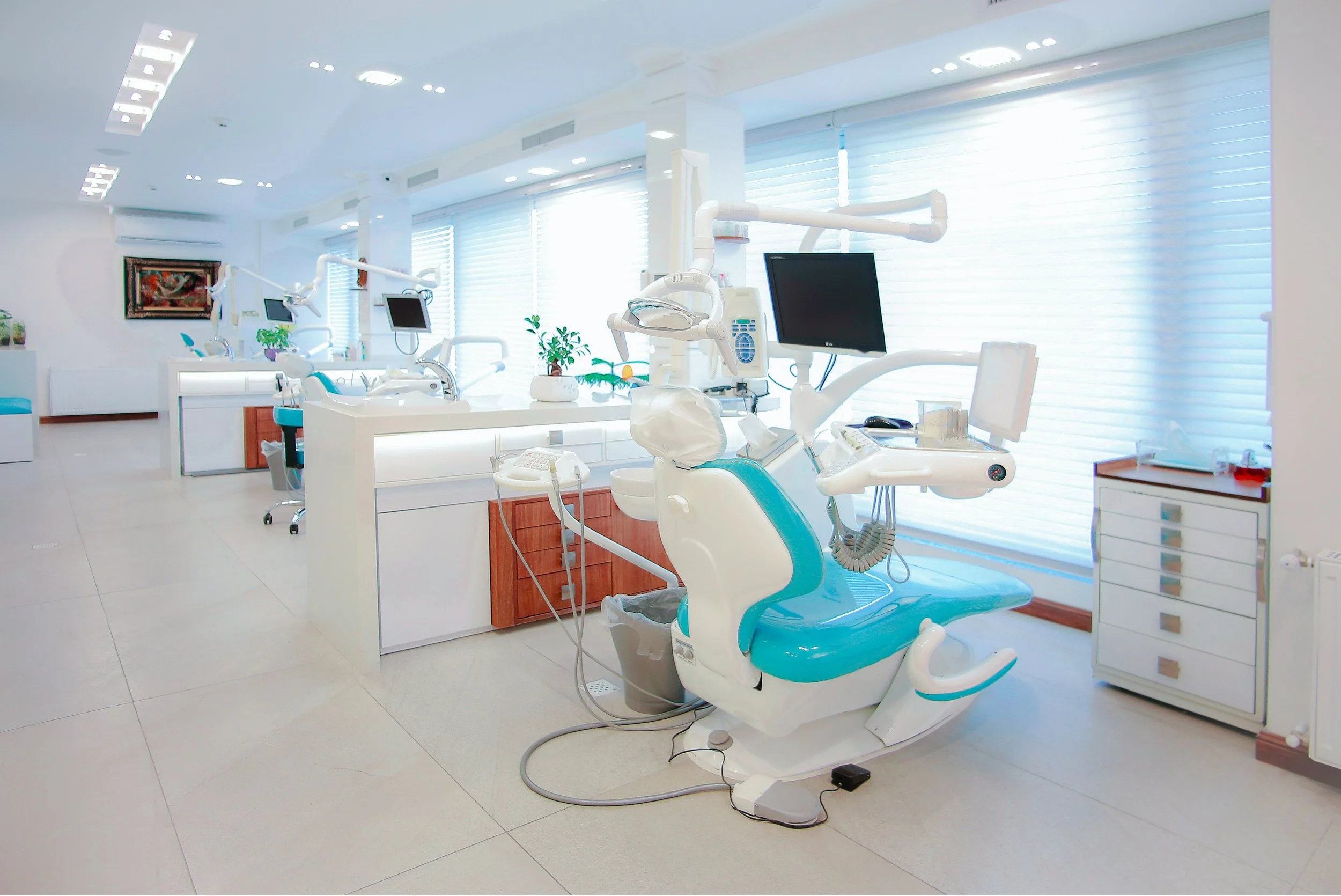 A modern dental clinic with dental chairs, monitors, and dental equipment in a bright, clean room with large windows and white walls.