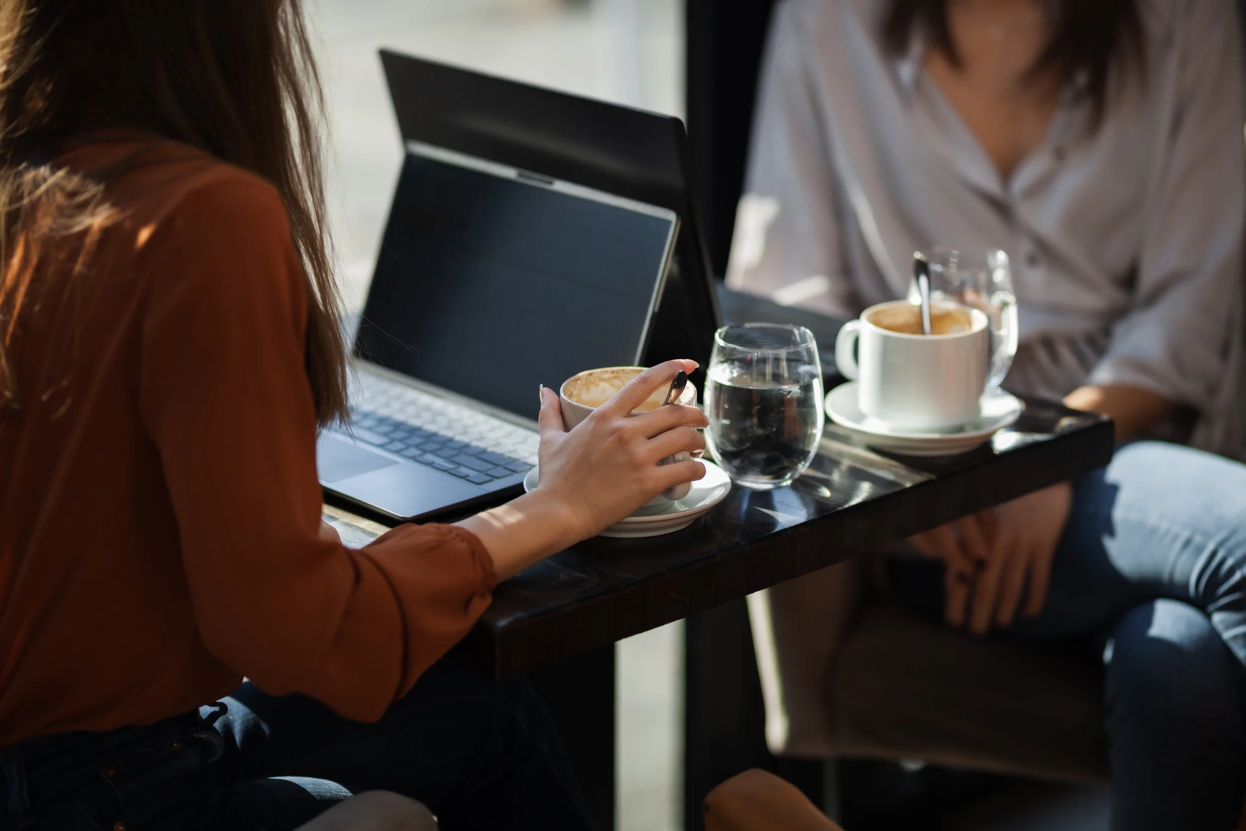 Two women sitting at a table in a café with laptops, coffee, and water glasses, engaging in conversation.