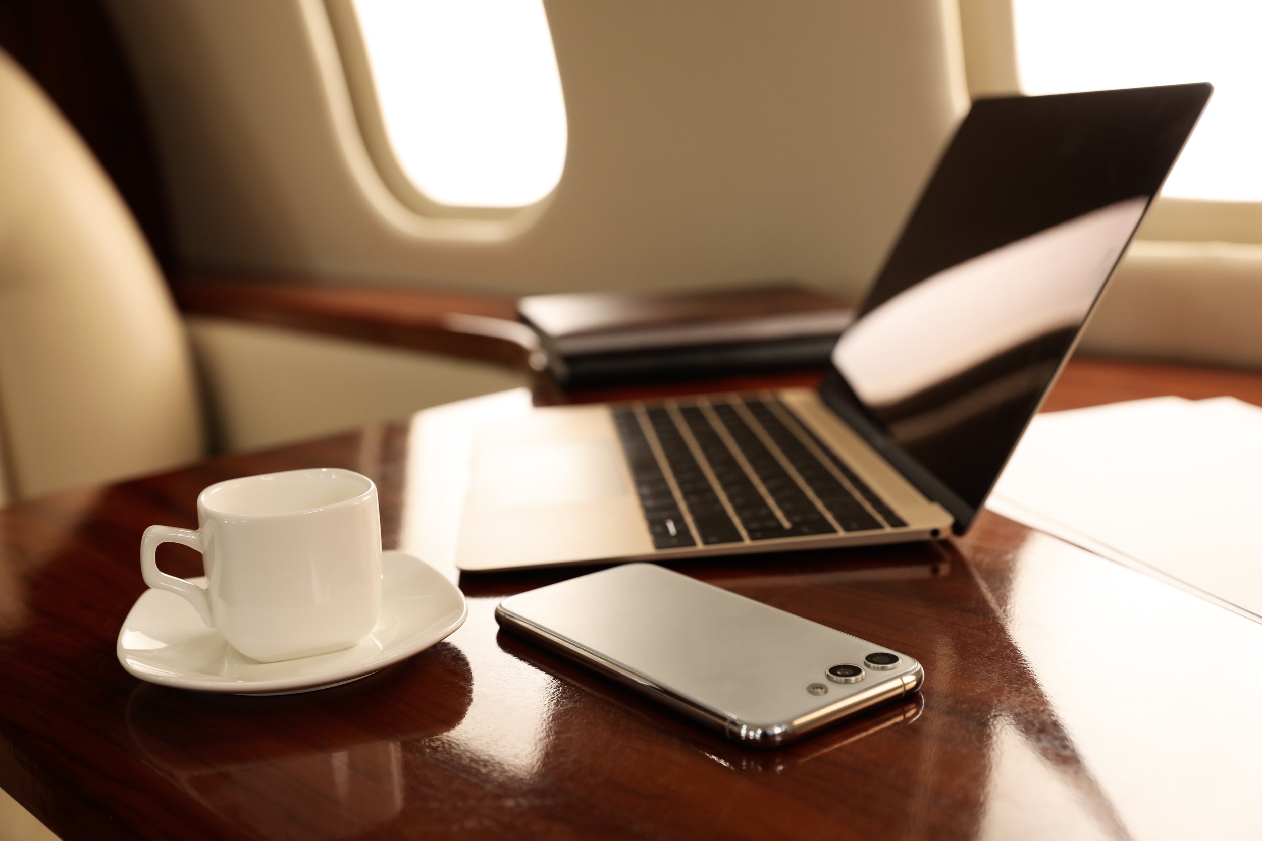 A table with a white coffee cup on a saucer, a laptop, a smartphone, and some papers, with an airplane window in the background.