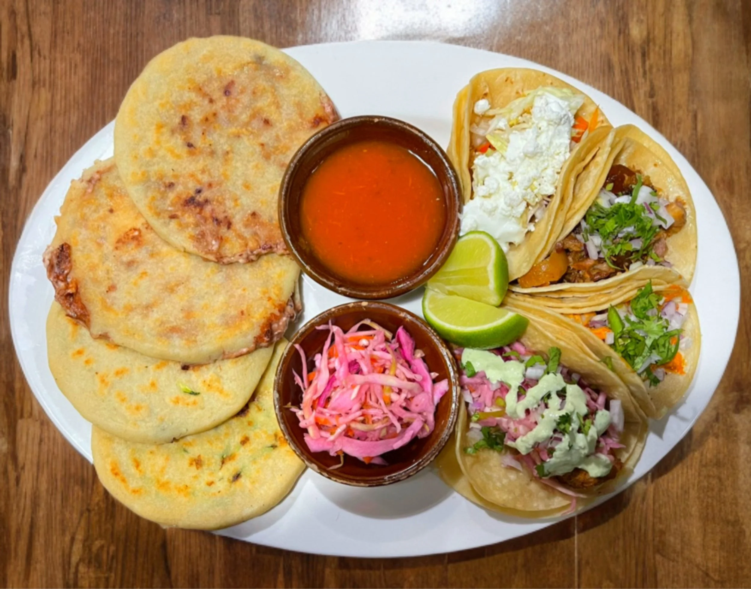Plate of assorted Mexican tacos with shredded lettuce, diced tomatoes, sour cream, cheese, and sliced lime, served with green and pink sauces.