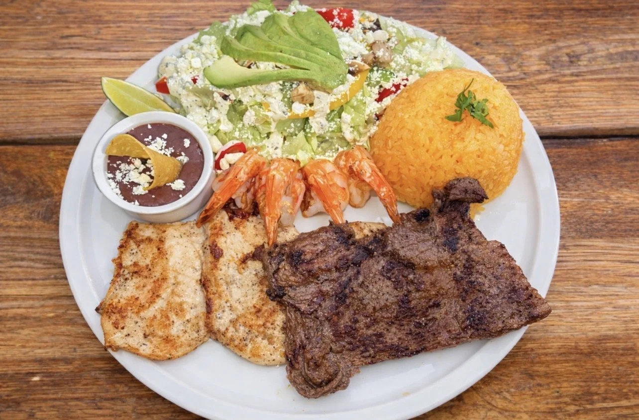 Plate of Mexican food with rice, salad, grilled meat, shrimp, refried beans, and lime wedge on a wooden table.