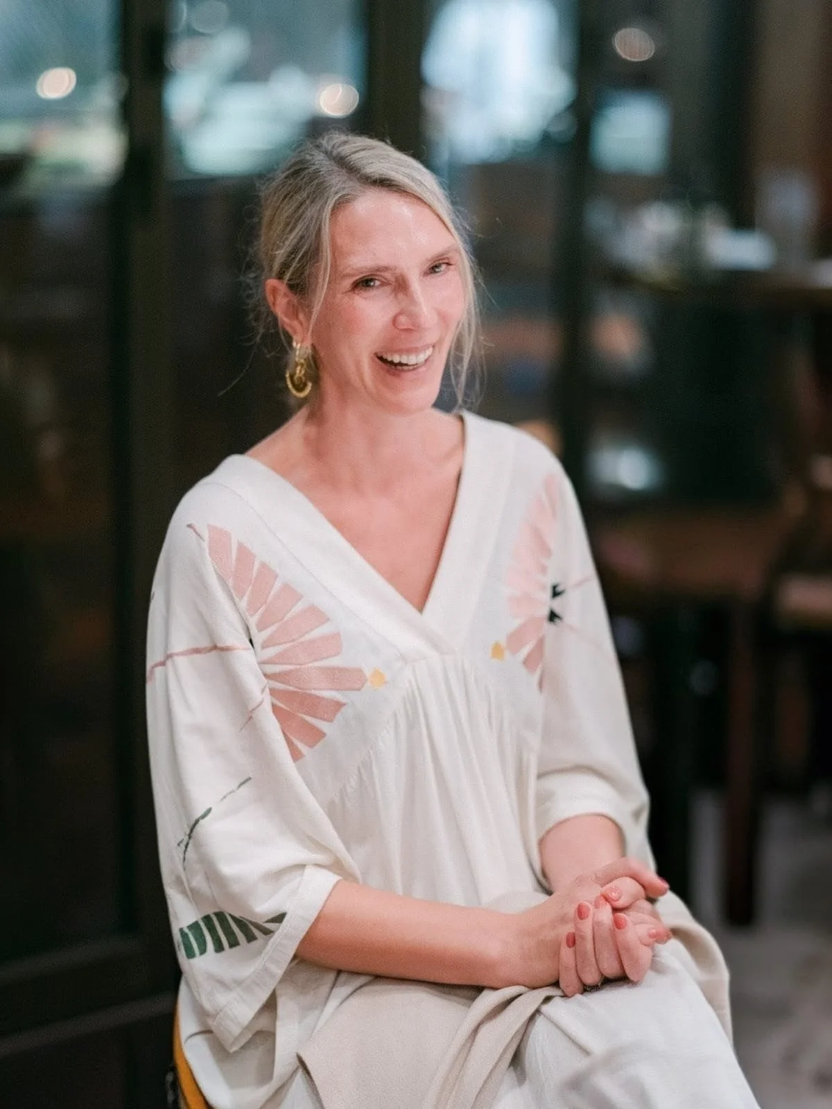 Smiling woman with blonde hair and hoop earrings, wearing a white dress with pink and green patterns, sitting with hands clasped in a casual indoor setting.