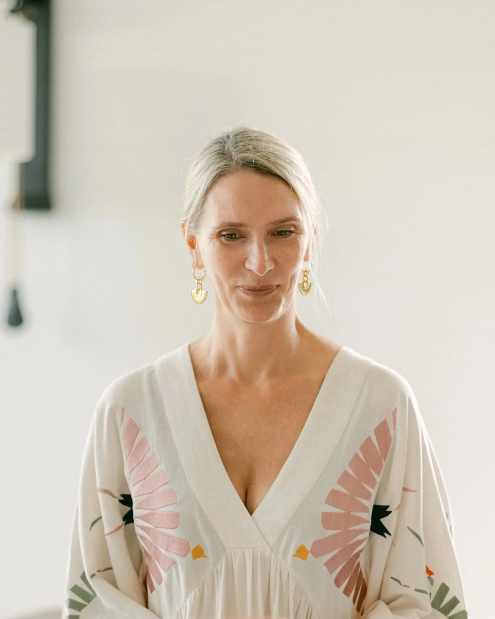 A woman with blonde hair and gold earrings is looking down and wearing a white dress with pink and orange leaf patterns.