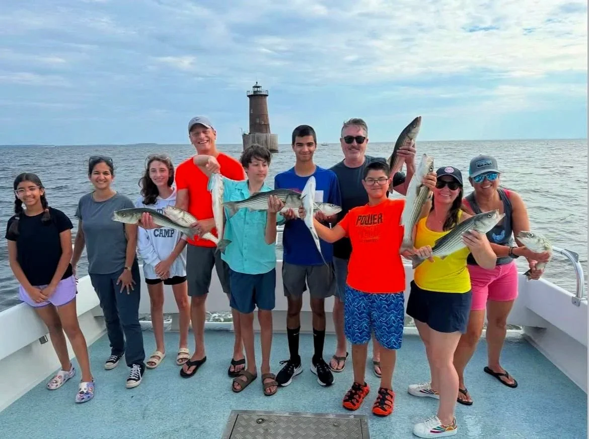 Group of people on a boat holding fish, with a lighthouse in the background at sea.