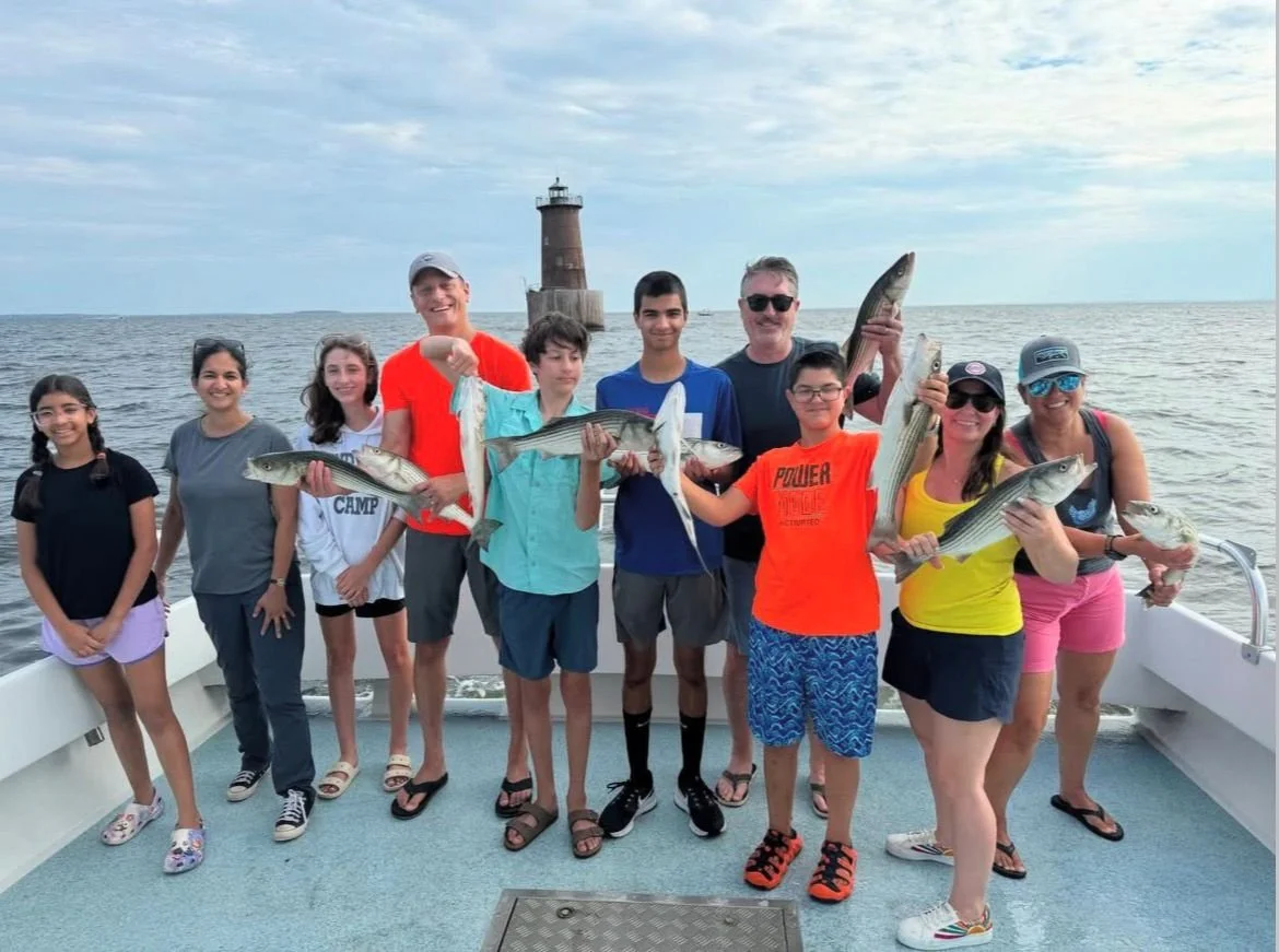 A group of people over ten, including children and adults, standing on a boat holding up fish they caught, with a lighthouse in the background on the water.