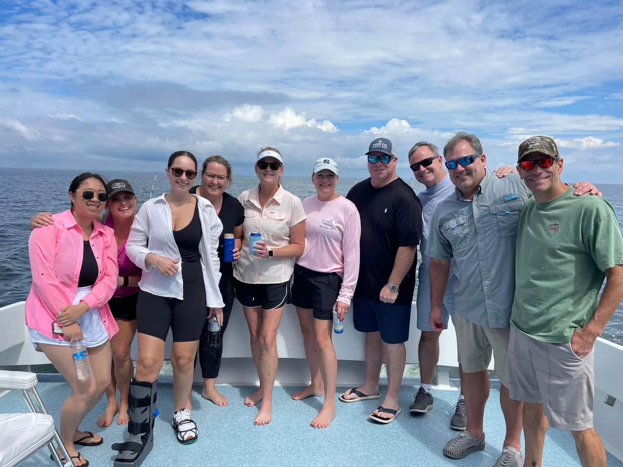A group of ten people, mostly women, standing together on a boat deck with the ocean and blue sky with scattered clouds in the background. Some are wearing sunglasses and casual summer clothing, and a few are holding water bottles.