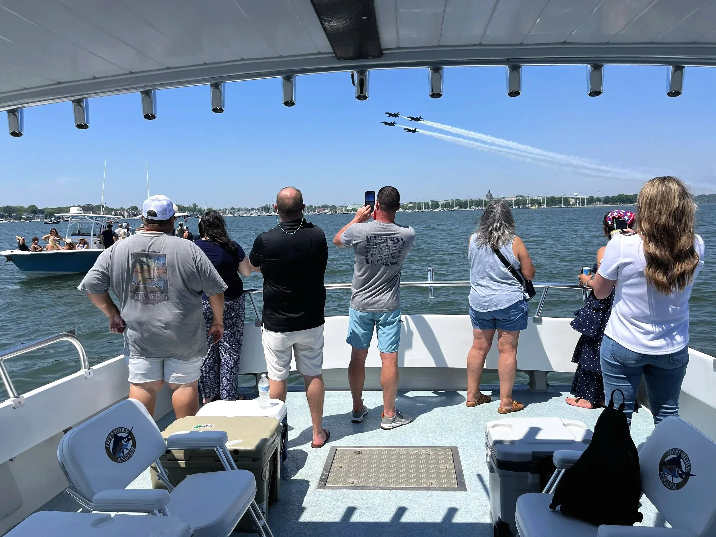 People on a boat watching an air show with jets flying overhead, leaving white trails in the blue sky.