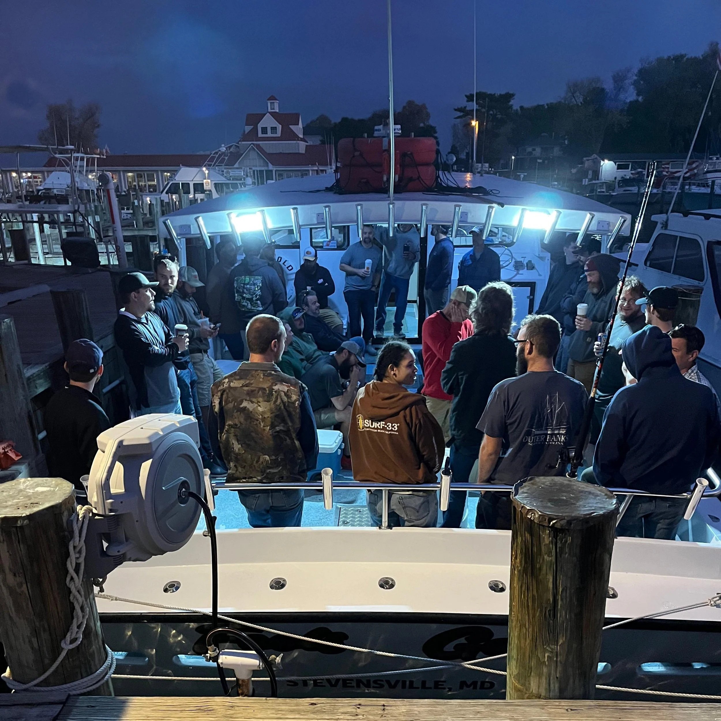 View from a dock showing a white boat in a marina during evening with a crowd of people on the deck, surrounded by wooden posts and ropes, with a building and trees in the background.
