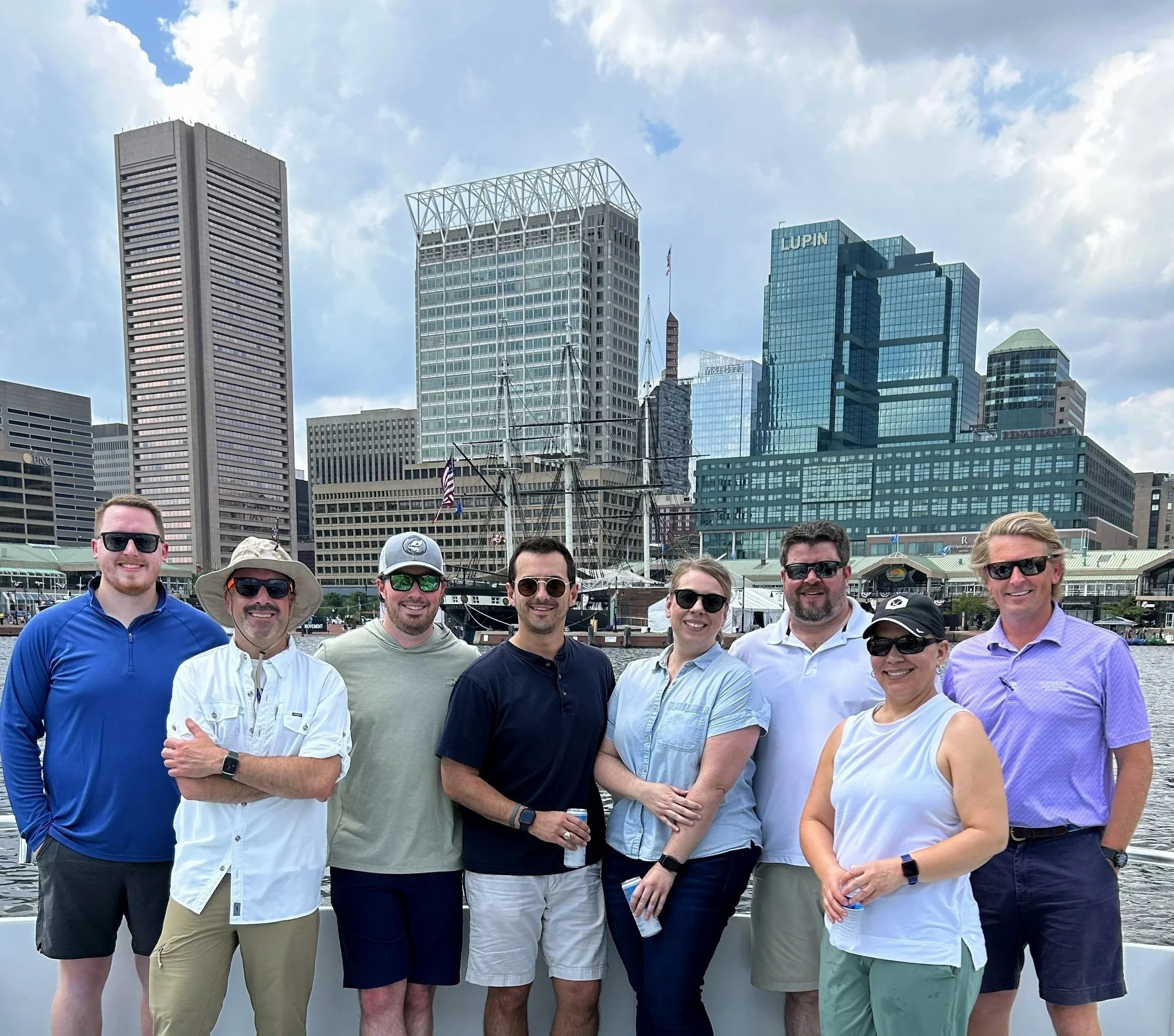 A group of people smiling and posing for a photo by the waterfront in front of tall city buildings, including ones labeled 'Lupin' and 'PNC', with a cloudy sky above.