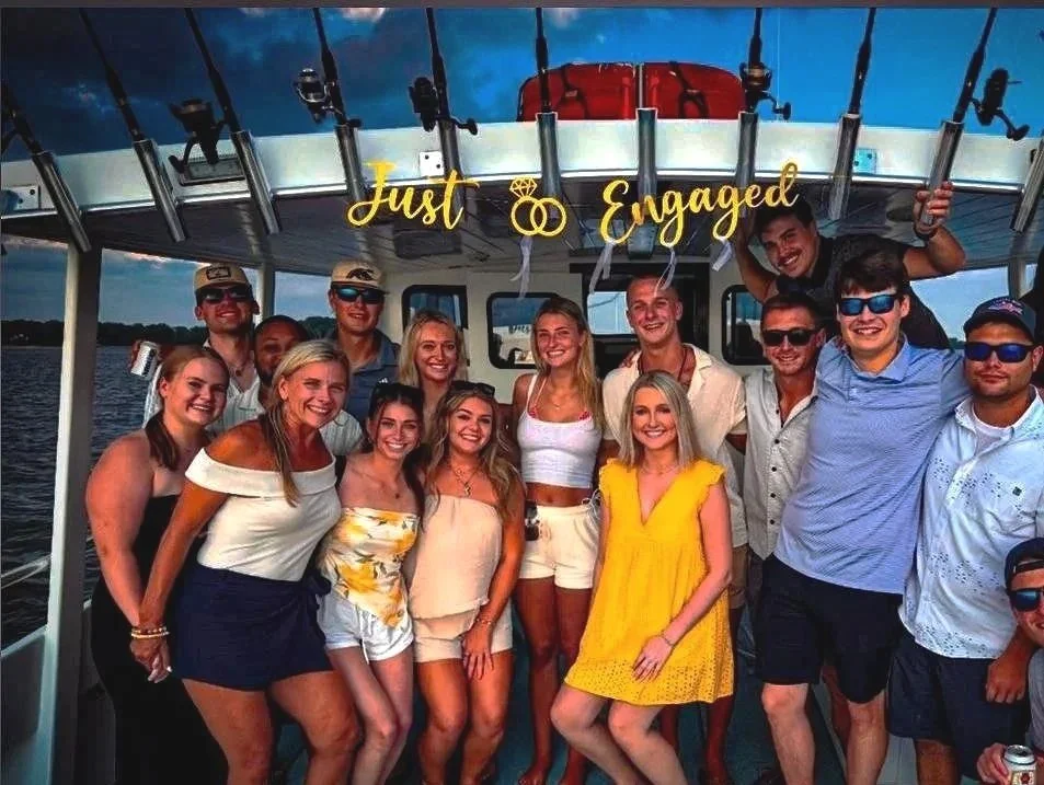 A group of friends on a boat celebrating an engagement, with a sign that reads "Just Engaged" hanging above.