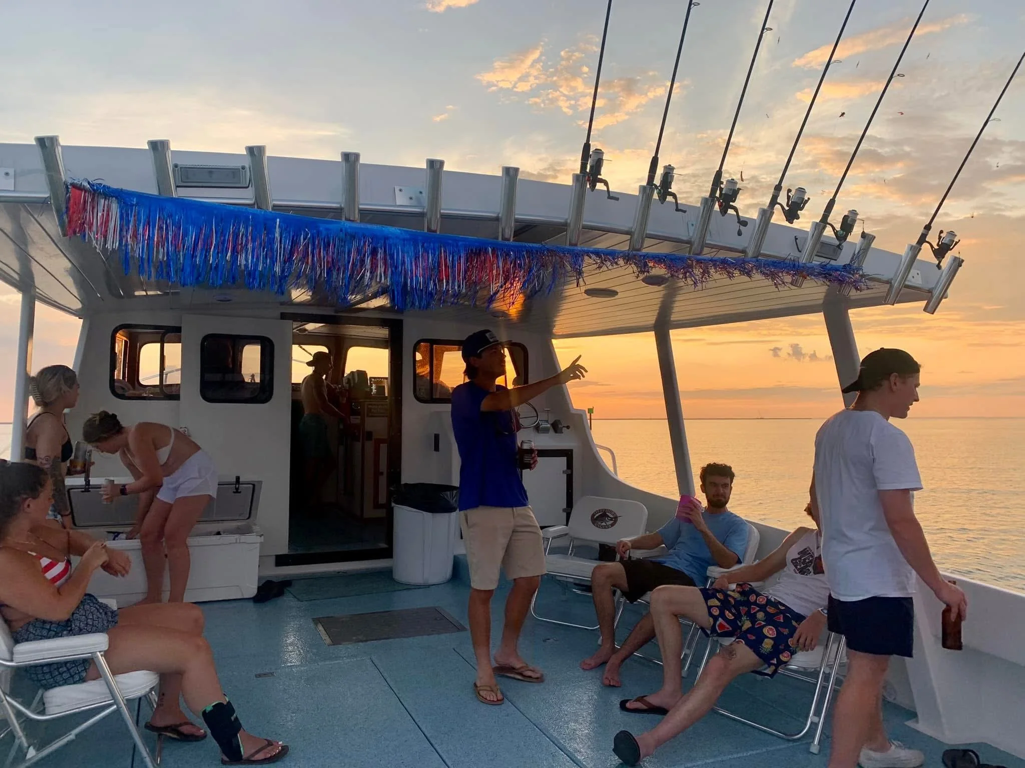 People socializing on a boat at sunset, with fishing rods on the upper deck and colorful decorations.