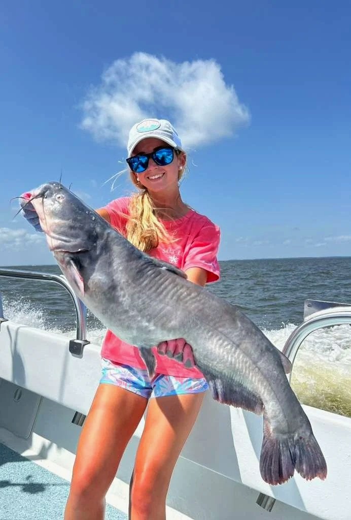 A woman on a boat holding a large fish with a cloud-shaped formation in the sky behind her.