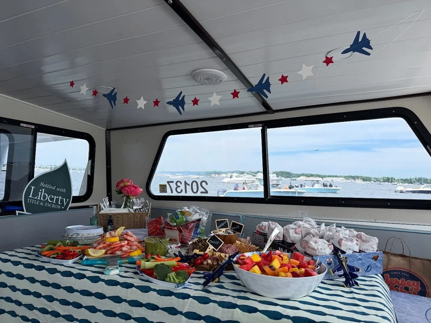 Table set for a celebration with various foods including fruit, vegetables, and bread on a boat, with a window showing water and boats outside, and patriotic decorations hanging from the ceiling.