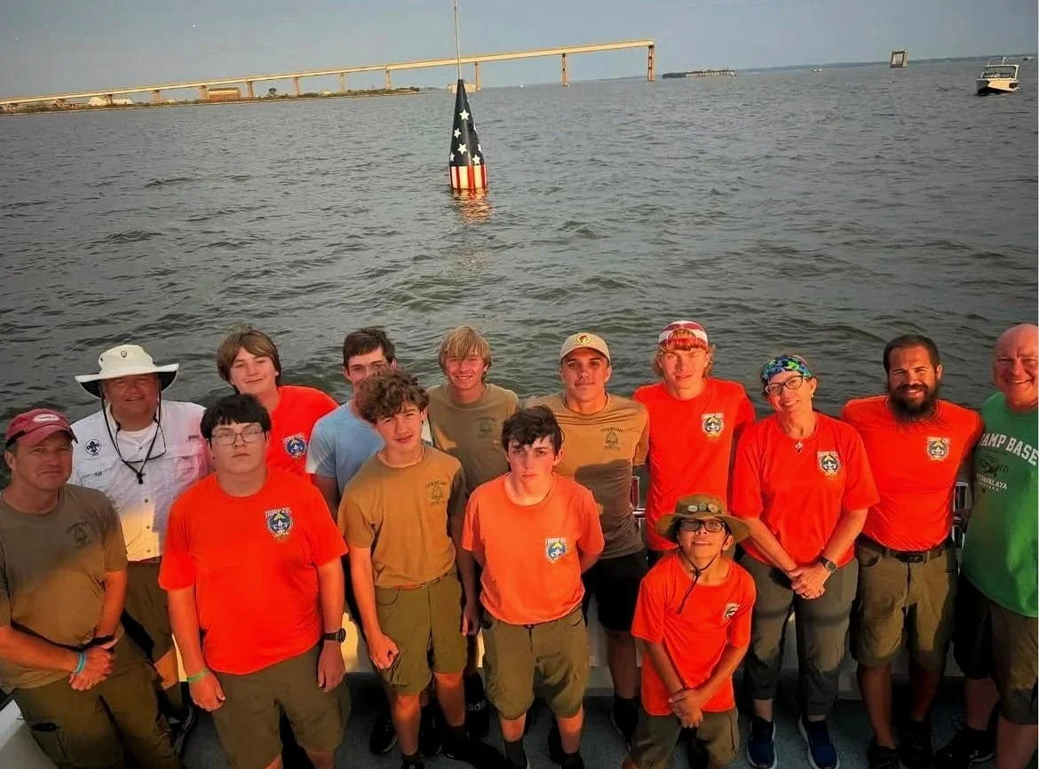 Group of diverse people, including children and adults, standing on a boat with a body of water and a bridge in the background during daytime.