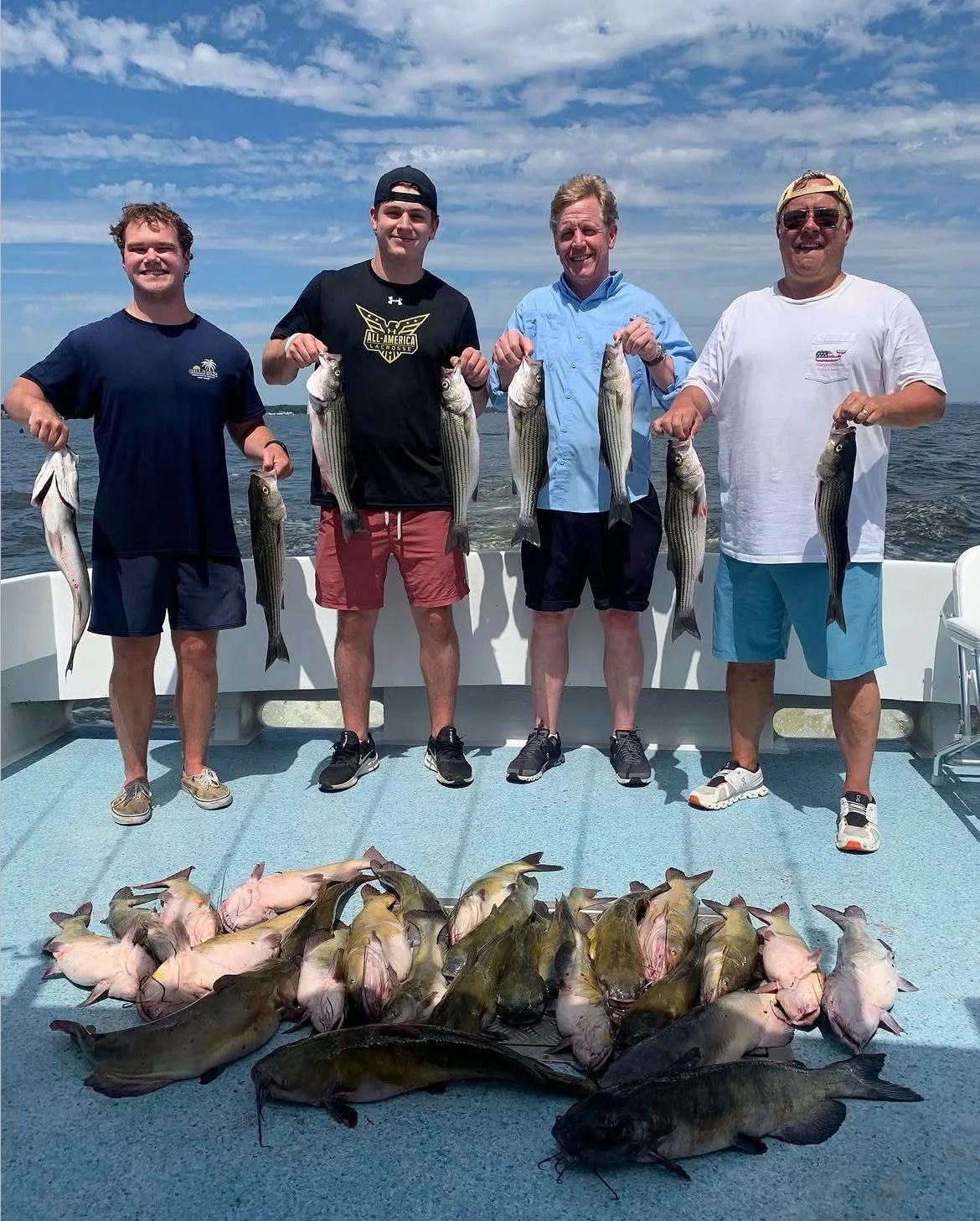 Four men standing on a boat holding caught fish with additional fish laid out on the boat's floor, with water and sky in the background.