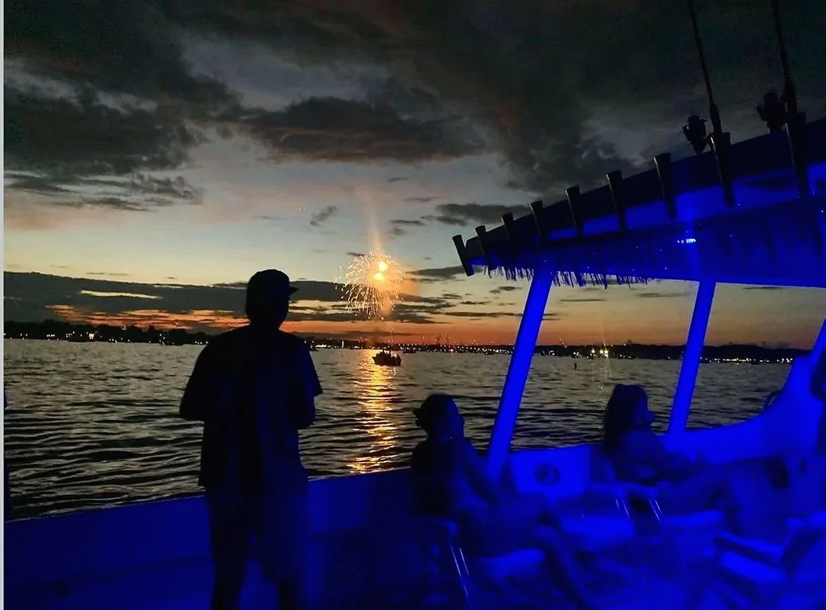 People on a boat watching fireworks over a body of water during sunset, with a cloudy sky and city lights in the distance.
