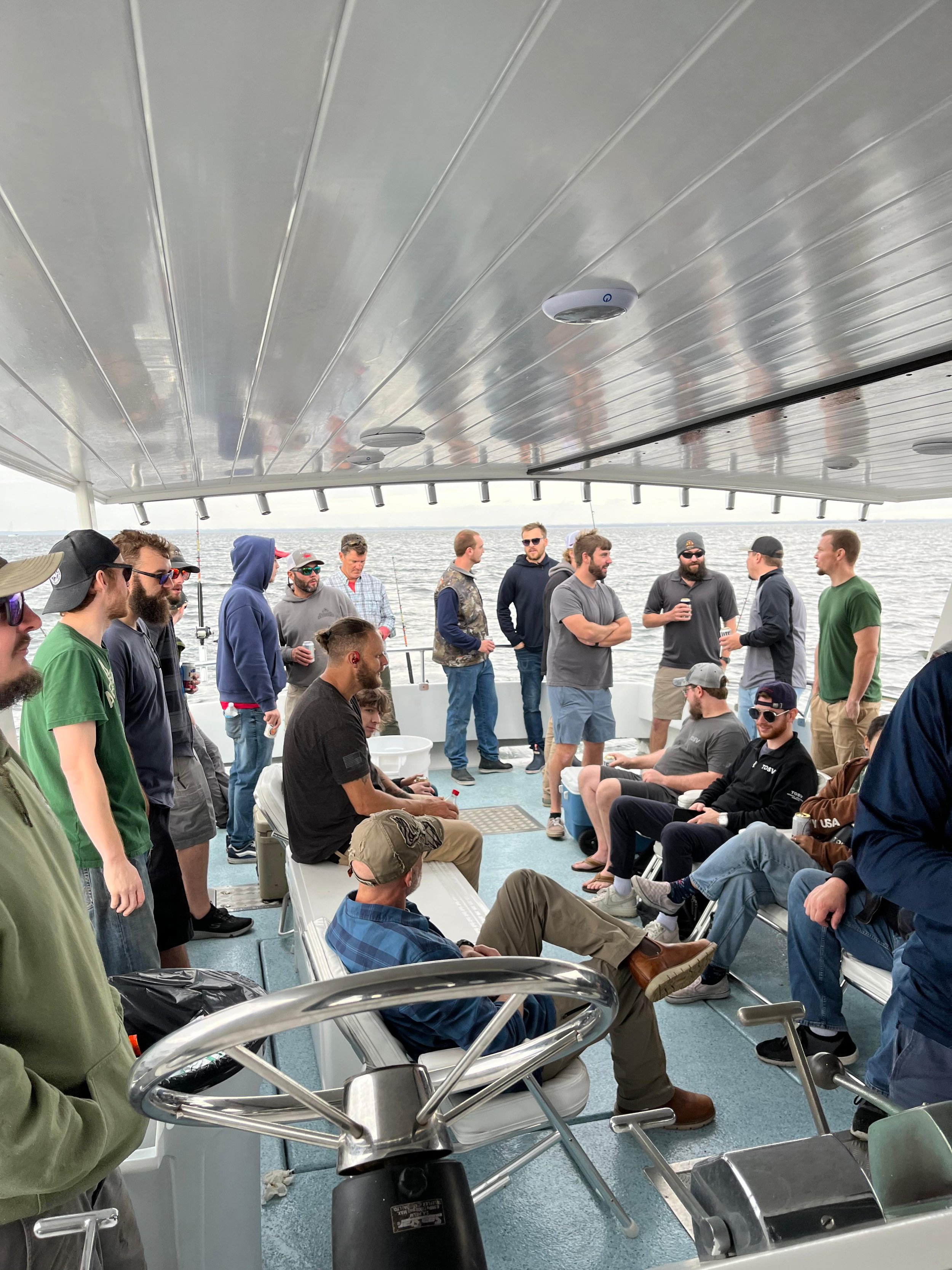 A group of people gathered on a boat deck, some sitting and some standing, enjoying a social event or conversation with water and a cloudy sky in the background.