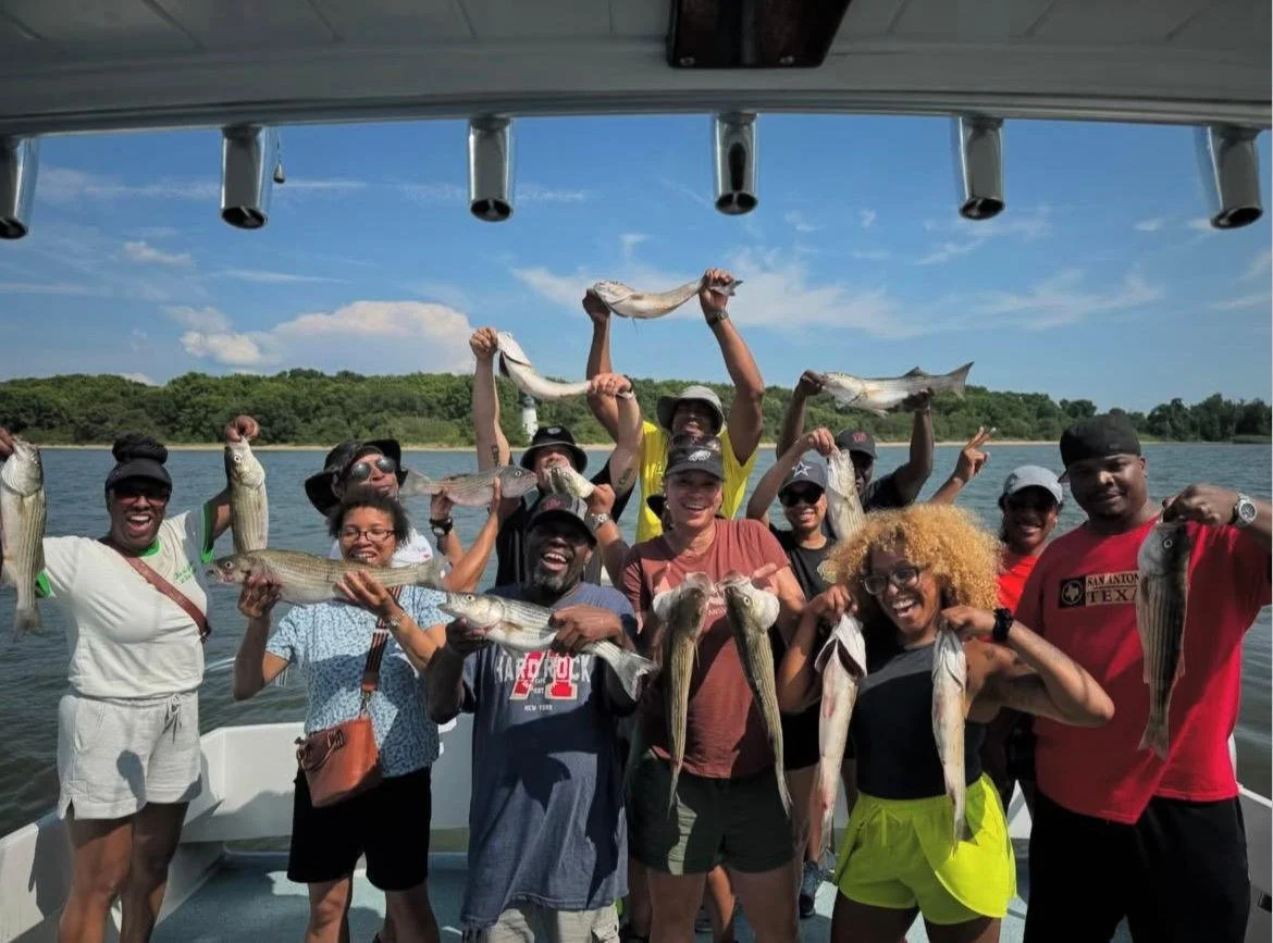 Group of people on a boat holding up fish they caught, smiling, with a water and tree-lined shoreline in the background.