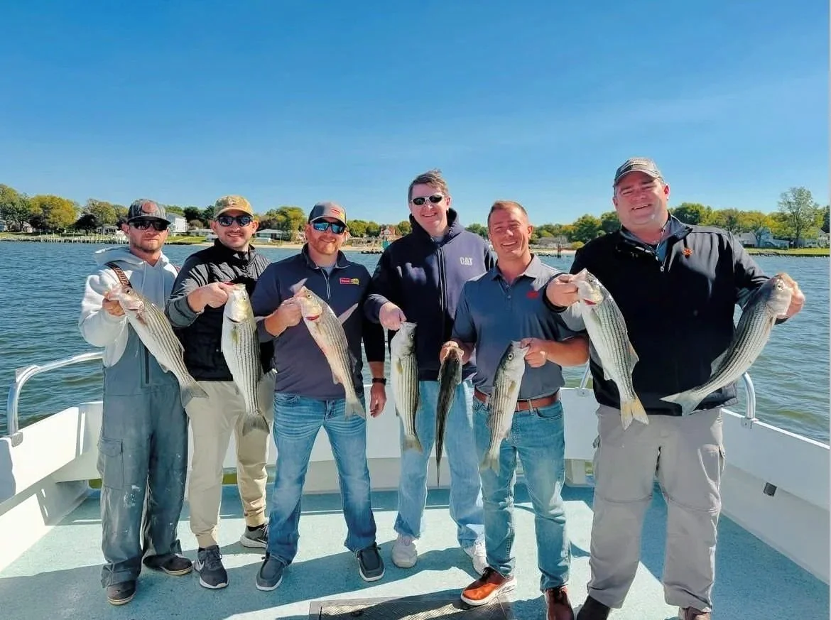 Six men on a boat holding fish they caught, with water and trees in the background.