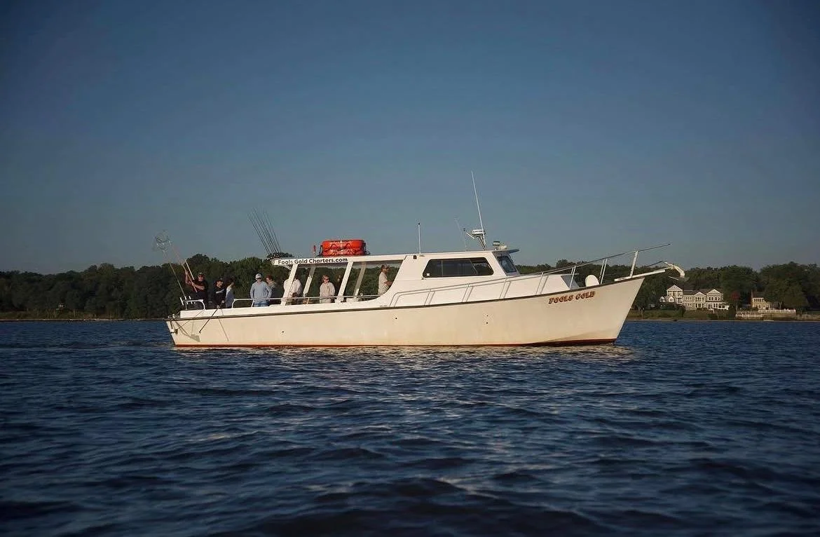 A white motor yacht named 'Fools Gold' floating on water with people on board, some fishing, and houses on the shoreline in the background.
