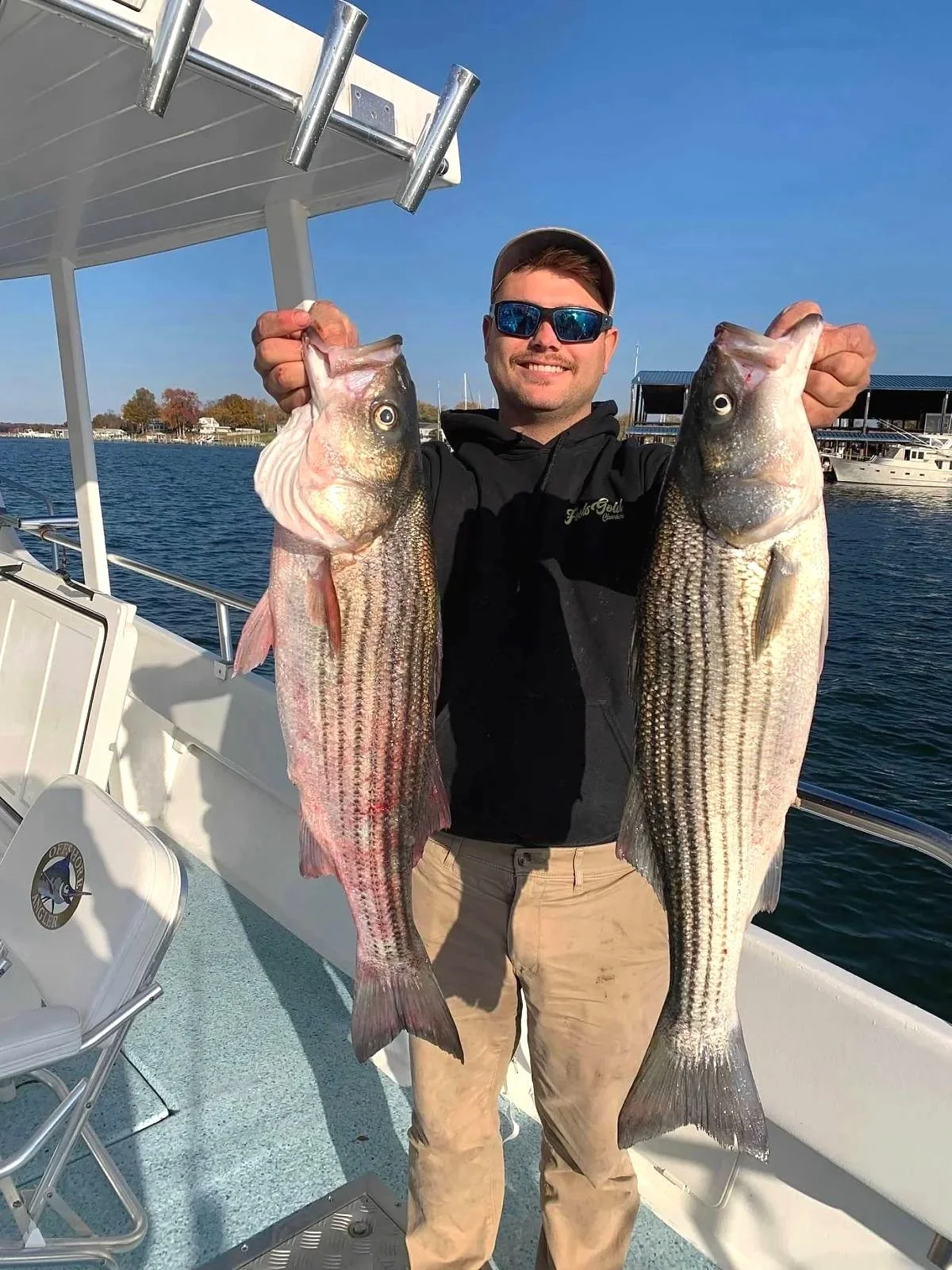Man on a boat holding two large striped bass fish, one in each hand, near the water with boats and buildings in the background on a clear day.