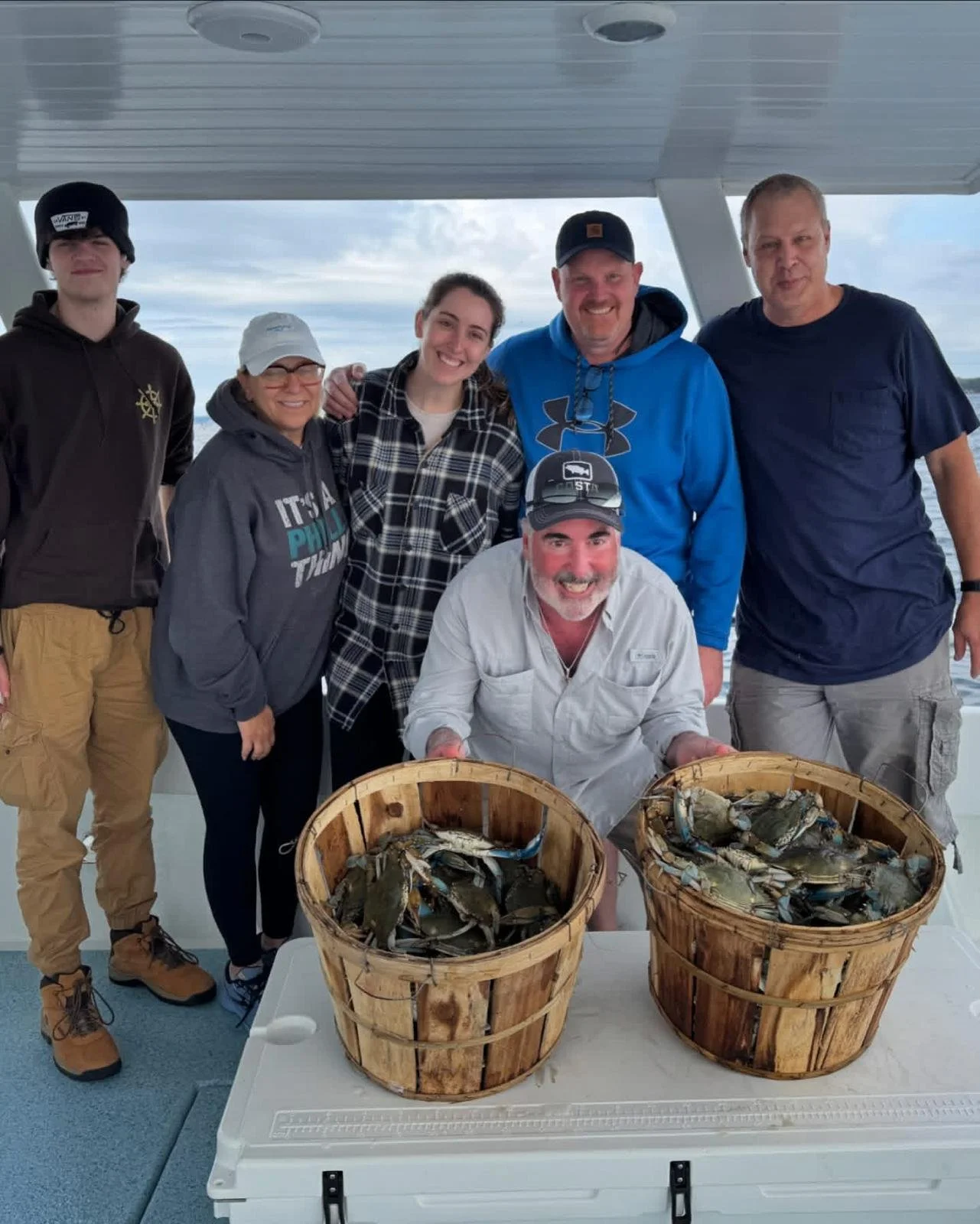 Group of seven people on a boat, two baskets filled with freshly caught fish or crabs, with water and cloudy sky in the background.