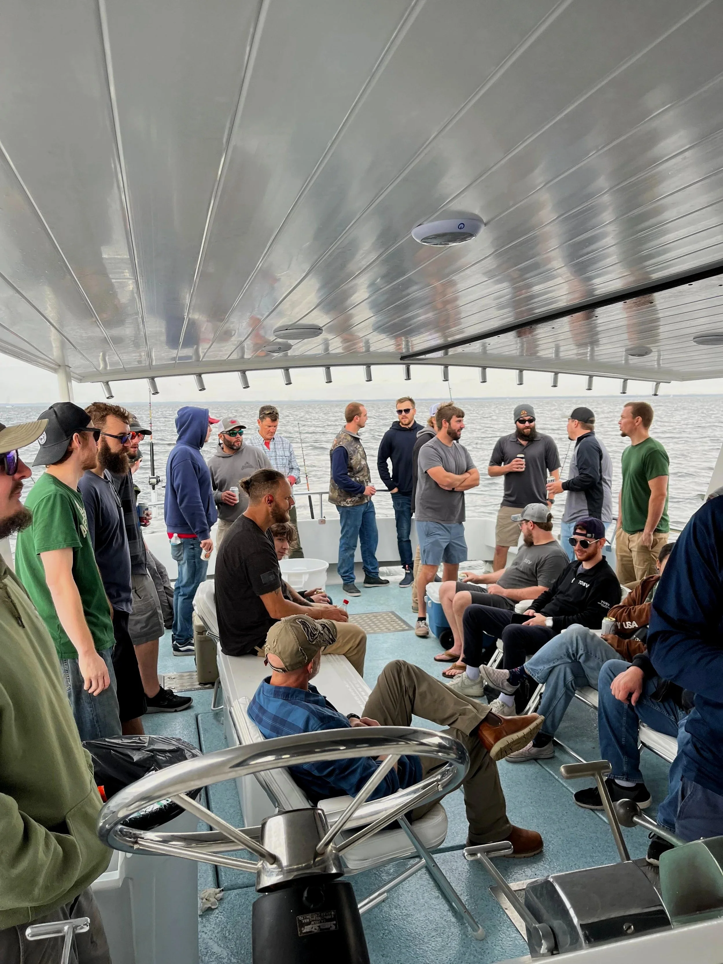A group of people gathered on a boat, some sitting and some standing, engaged in conversation, with the water visible in the background. The boat has a modern ceiling with built-in lights.