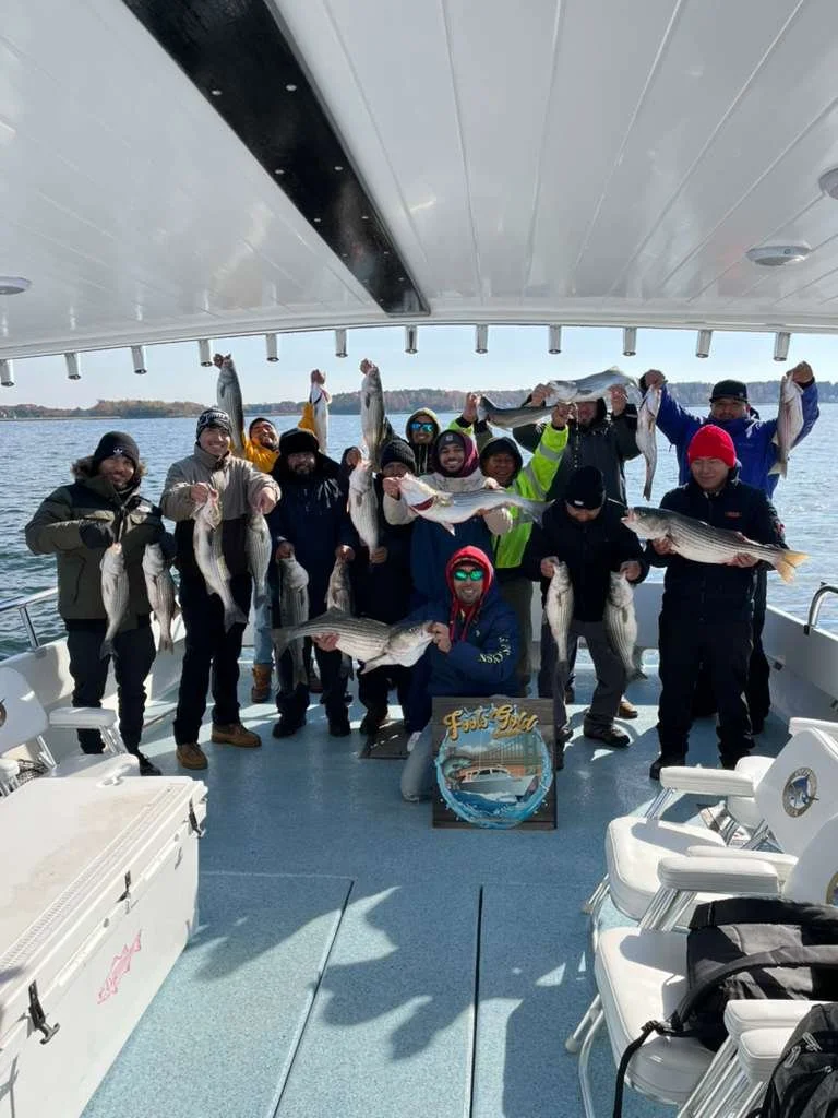 Group of people on a boat holding large fish they caught, with water and trees in the background.