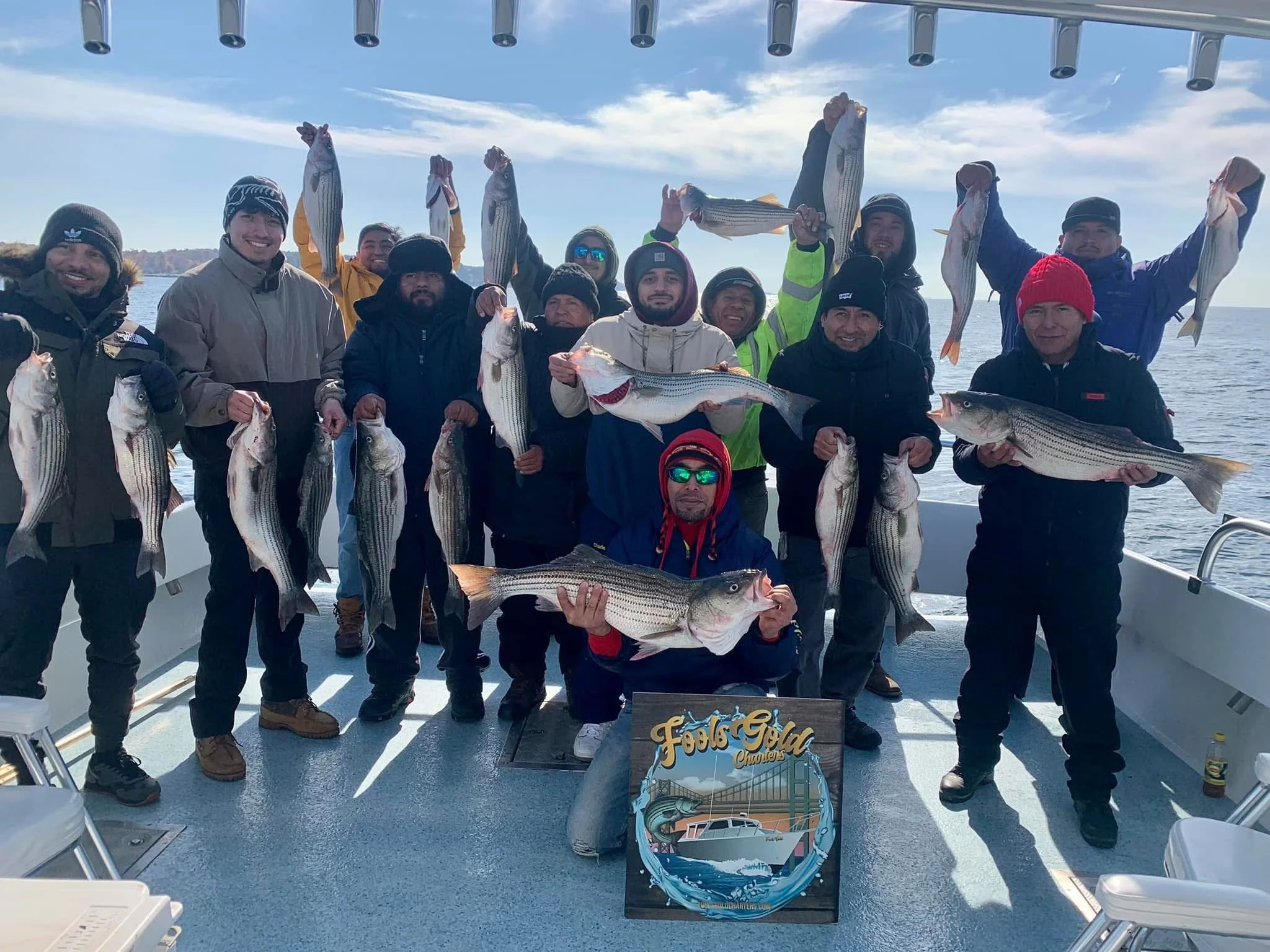 A group of people on a boat holding up large striped fish caught during a fishing trip, with a clear blue sky and water in the background.