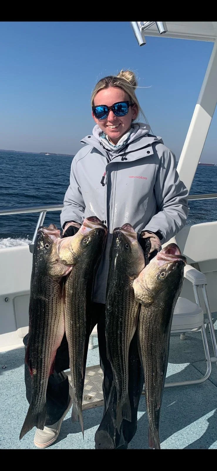 Woman wearing sunglasses and a light gray jacket on a boat, holding four large fish with the ocean in the background.