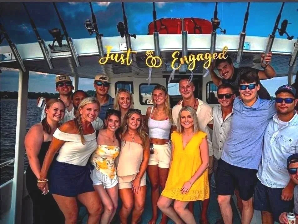Group of young adults on a boat celebrating an engagement. The sign above reads 'Just Engaged.' Everyone is smiling, some holding drinks, with a water backdrop.