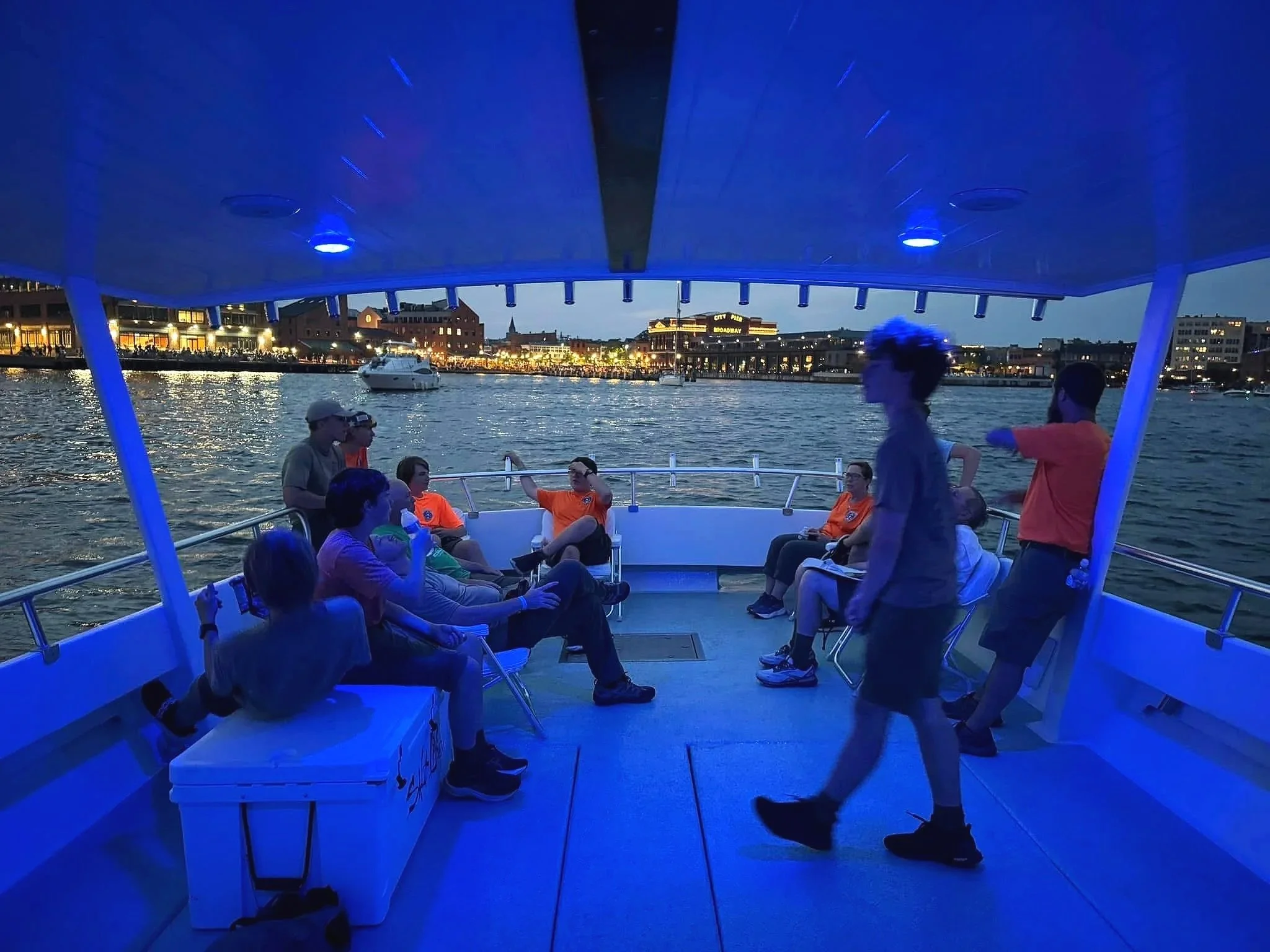 People relaxing and socializing on a boat at dusk, with city buildings and boats visible across the water in the background.