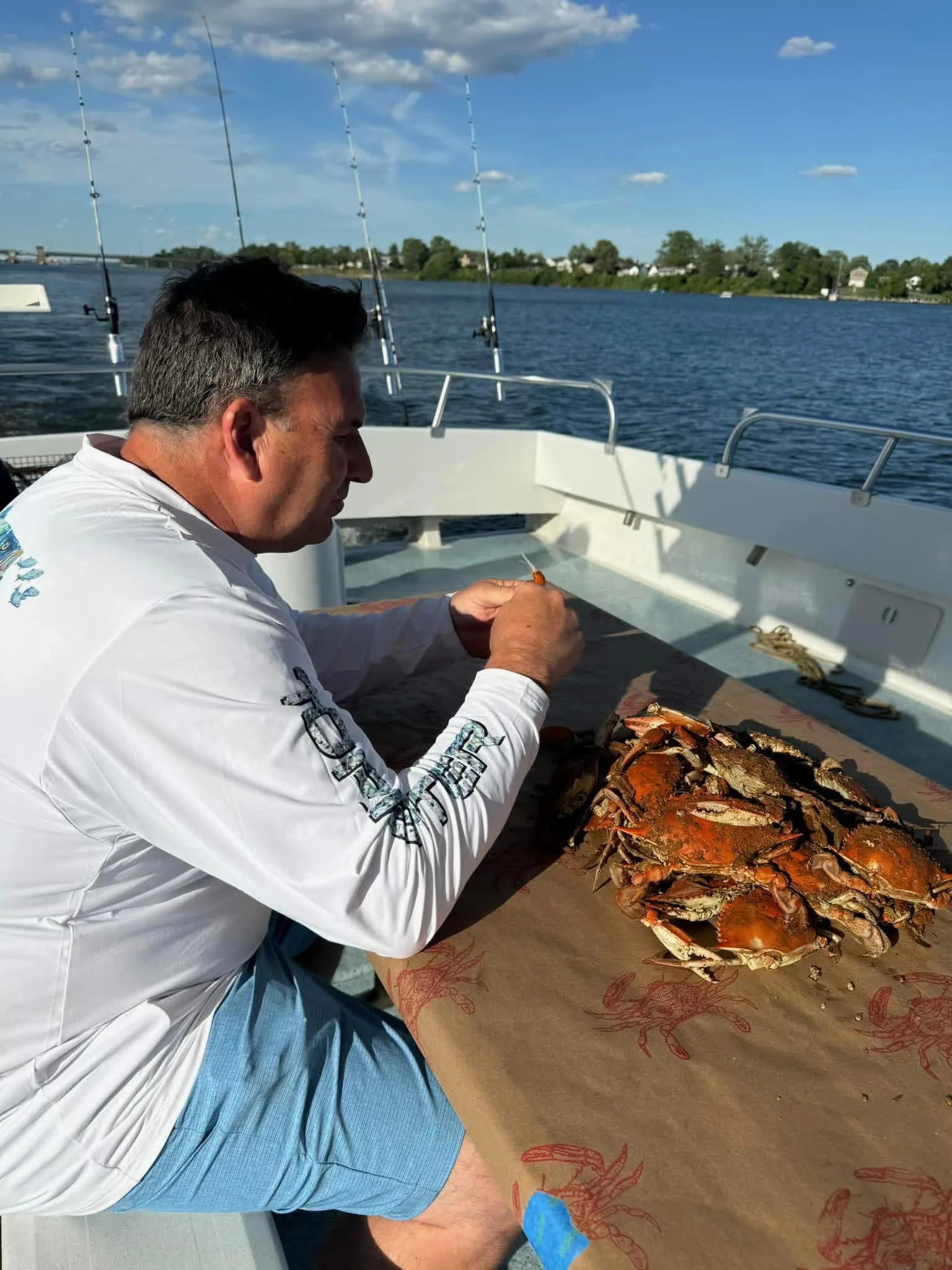 A man sitting on a boat by the water preparing to cook a pile of cooked crabs on a table covered with lobster-print paper, with fishing rods visible in the background, and a shoreline with houses and trees under a partly cloudy sky.