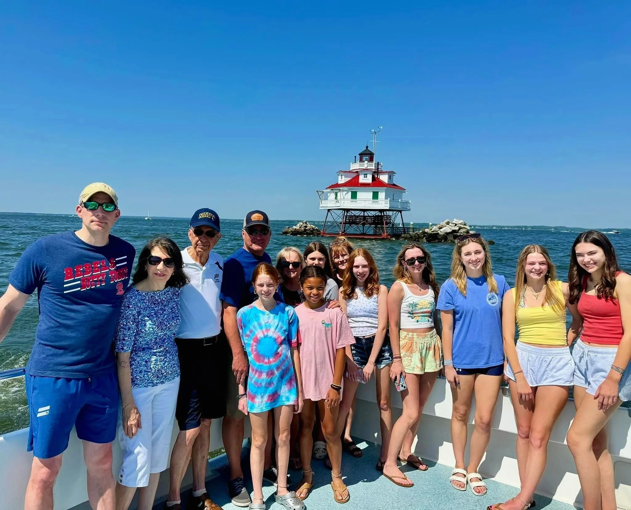 Group of people standing on a boat deck near a lighthouse with the ocean in the background on a sunny day.