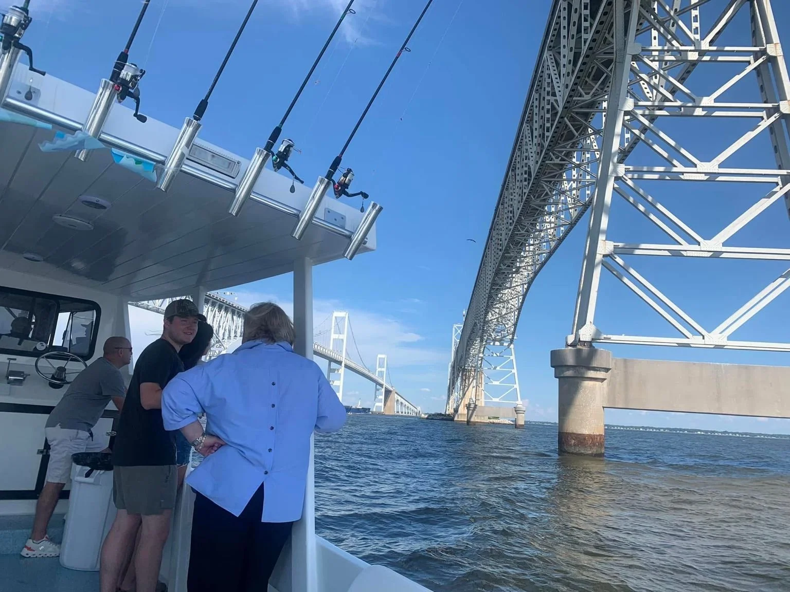 People on a boat with fishing rods under a clear blue sky, with large bridges in the background over the water.