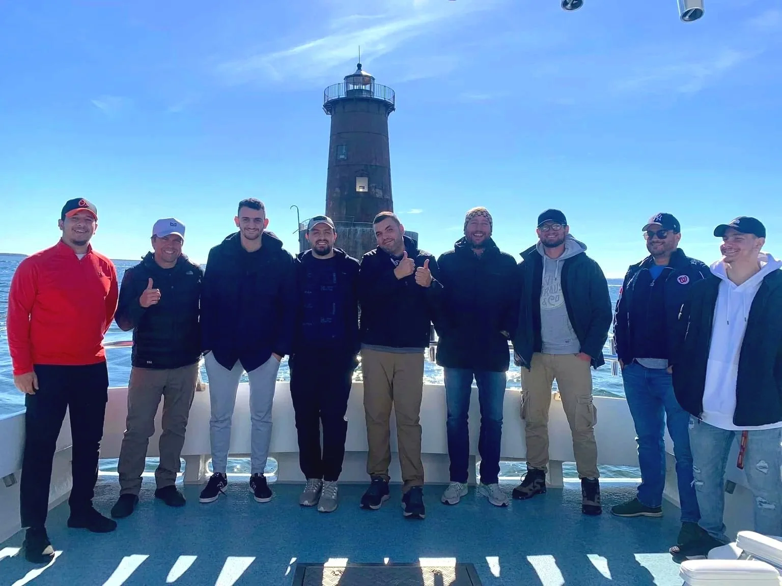 A group of ten young men standing on a boat deck with a lighthouse in the background, blurry sky and water visible, some smiling and giving thumbs up.