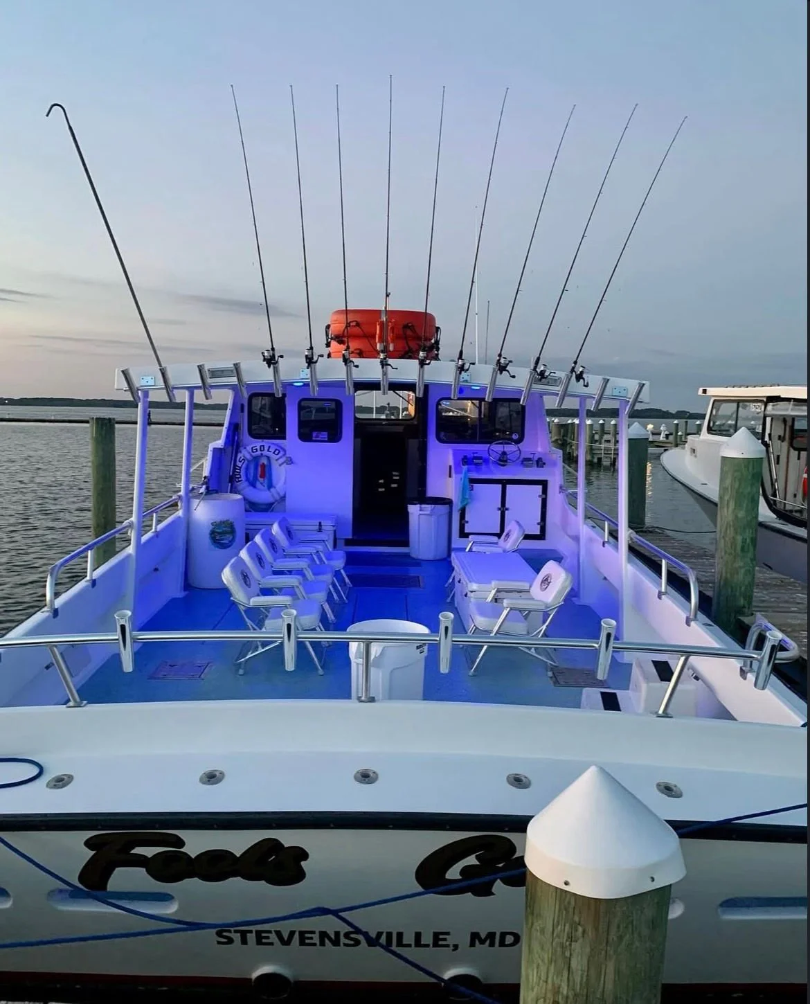A white boat docked at a marina with multiple fishing rods mounted on the upper deck, illuminated with blue lighting, and with chairs arranged on the deck.