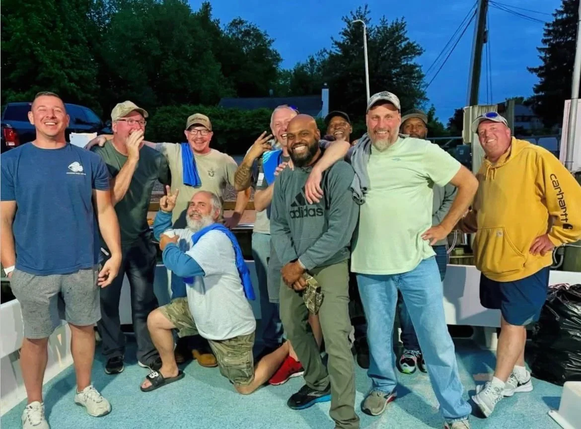 Group of men smiling and posing together outdoors during evening, standing on a boat or dock, with trees and vehicles visible in the background.