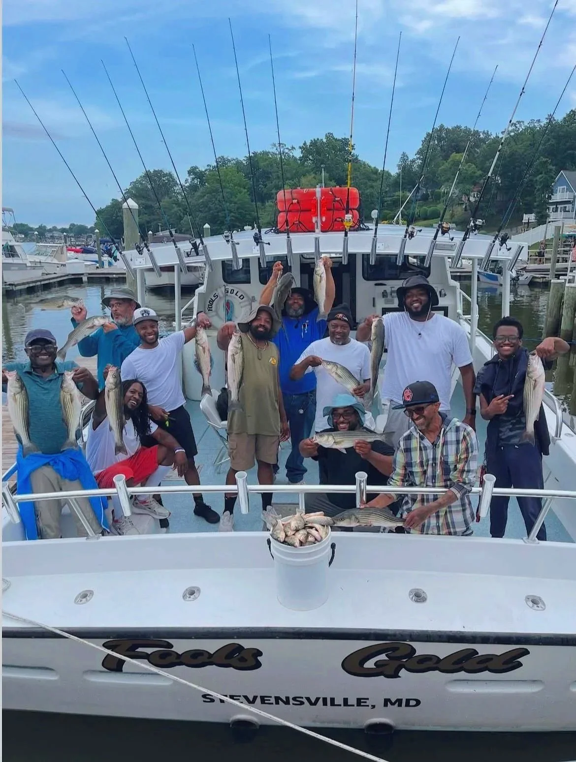 Group of people on a boat at a marina, holding caught fish and smiling, with water and other boats in the background.